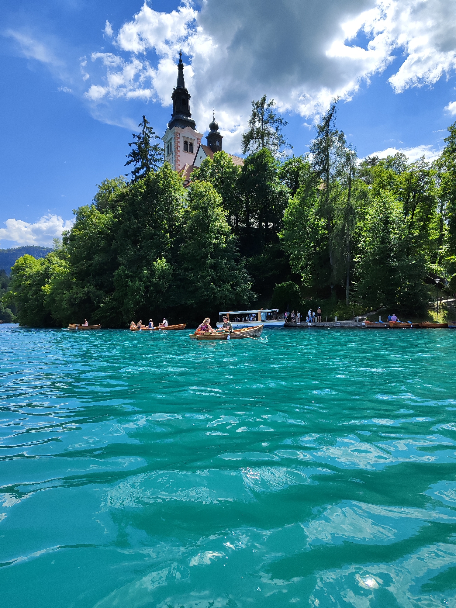 This image depicts a view of Lake Bled in Slovenia, looking towards the Church of the Assumption on Bled Island. The foreground is dominated by the vibrant turquoise waters of the lake, with ripples and reflections of the sky visible. Several wooden rowboats are scattered across the lake, some occupied by people. In the middle ground, a larger white boat with a canopy is docked near the shore, with several people standing on a wooden platform beside it. The background features a lush green forest that covers the island and surrounding hills, with the Church of the Assumption prominently visible atop the island, its steeple reaching towards a sky filled with white, fluffy clouds. The overall scene conveys a serene and picturesque summer day.