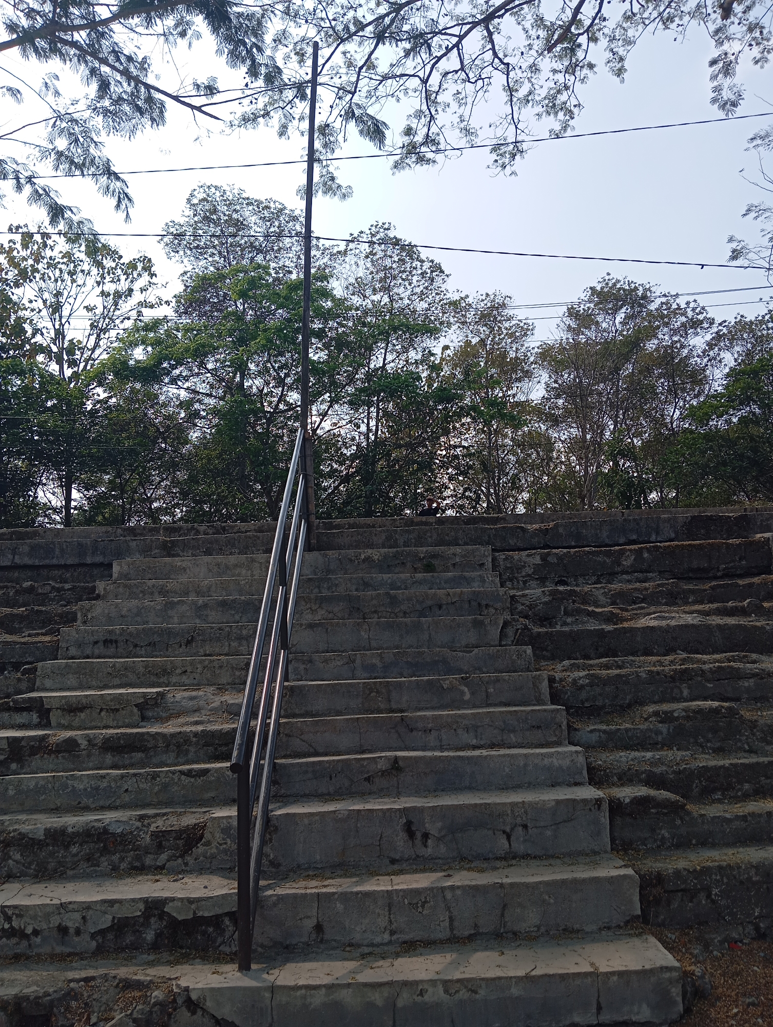 The image shows a concrete staircase with a metal railing, leading upwards. The staircase appears to be outdoors, with trees and vegetation visible in the background. The sky is overcast, and there are power lines running across the upper part of the image. The concrete of the stairs is weathered and cracked in places, suggesting age and exposure to the elements. There is some debris, such as small stones and leaves, scattered on the steps. The scene is static and does not depict any activity or interaction. The location is identified as Sokaraja, Indonesia.