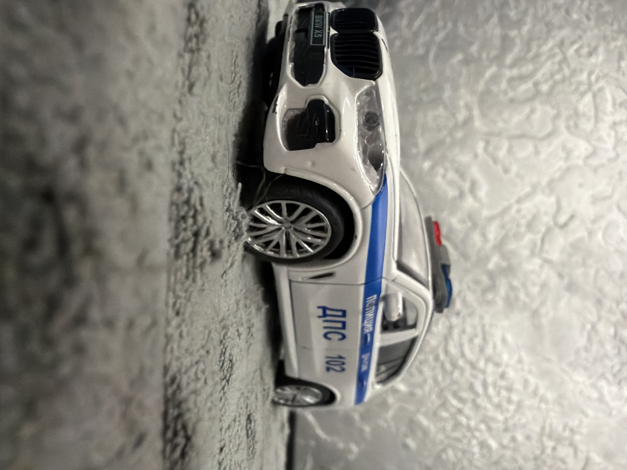 A white BMW X5 police car toy is positioned on a gray, fuzzy surface, likely a carpet or rug. The toy car is facing right, with its front right wheel slightly turned. It has a blue stripe running along its side, and Russian text which reads "ПОЛИЦИЯ" (Police) and "ДПС 102" (Road Patrol Service 102). The car has a visible license plate with "BMW X5" written on it. A red and blue emergency light bar is mounted on the roof. The background is a textured, light-colored surface. There is no indication of Mytishchi, Russia, or any specific urban environment in the image.