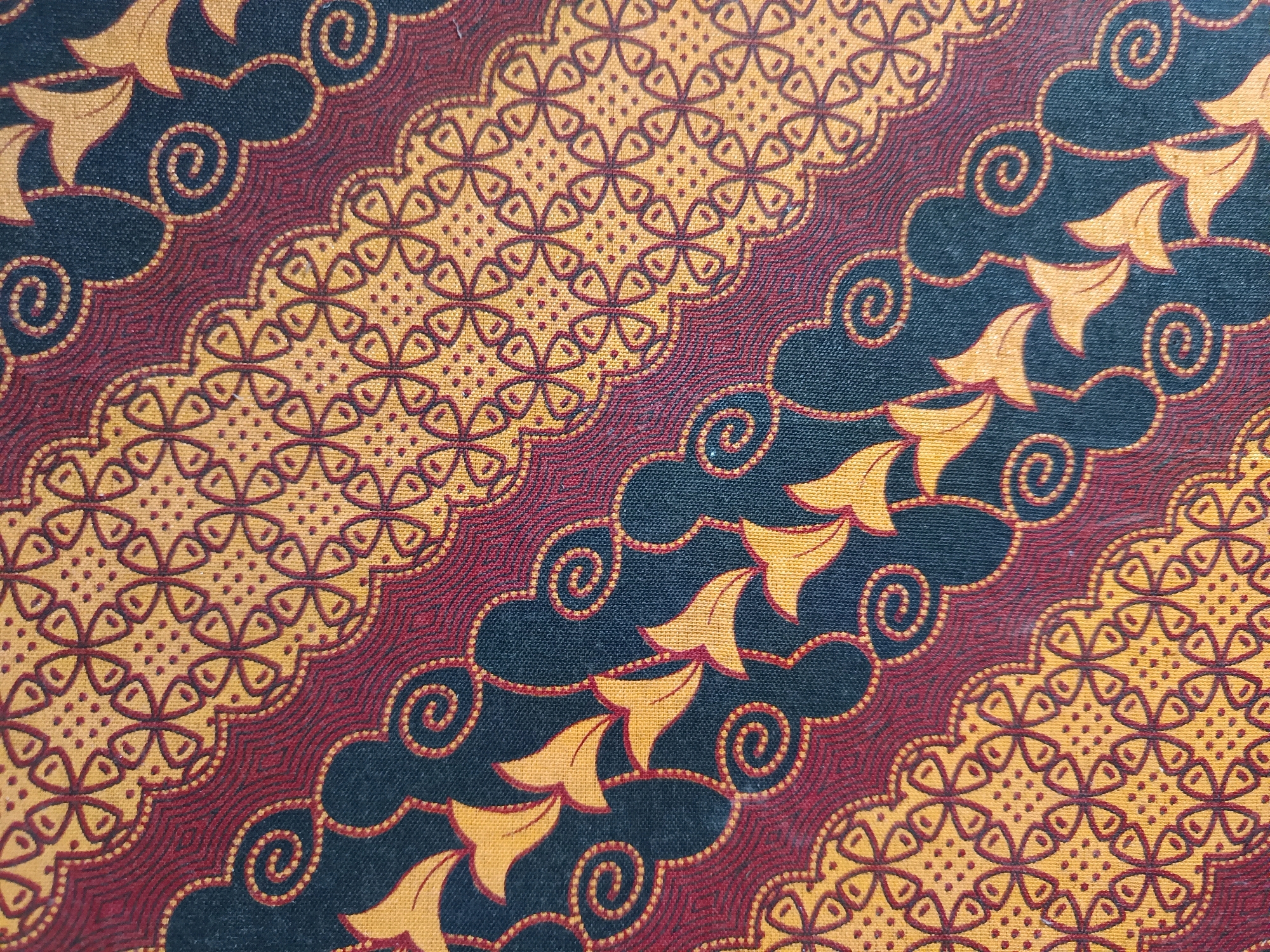 This image is a close-up, overhead view of a batik fabric with a dark blue background. The fabric features a repeating pattern of stylized floral motifs and swirling lines in shades of yellow, gold, and deep red. The pattern is diagonal, with wavy borders separating different sections. In one section, there are repeating motifs that resemble stylized flowers or butterflies, each with four petal-like shapes and filled with a dot pattern. Adjacent to this, a band of red and gold wavy lines is visible. Further across the fabric, there are triangular, leaf-like shapes in yellow, outlined in red, interspersed with red and gold spiral designs. The texture of the fabric appears to be cotton, with a visible weave. The lighting is even, suggesting it was taken in a well-lit indoor environment or outdoors under diffused light. There are no people or animals visible in the image, and no context is provided to place it in Gunung Sekar, Indonesia.