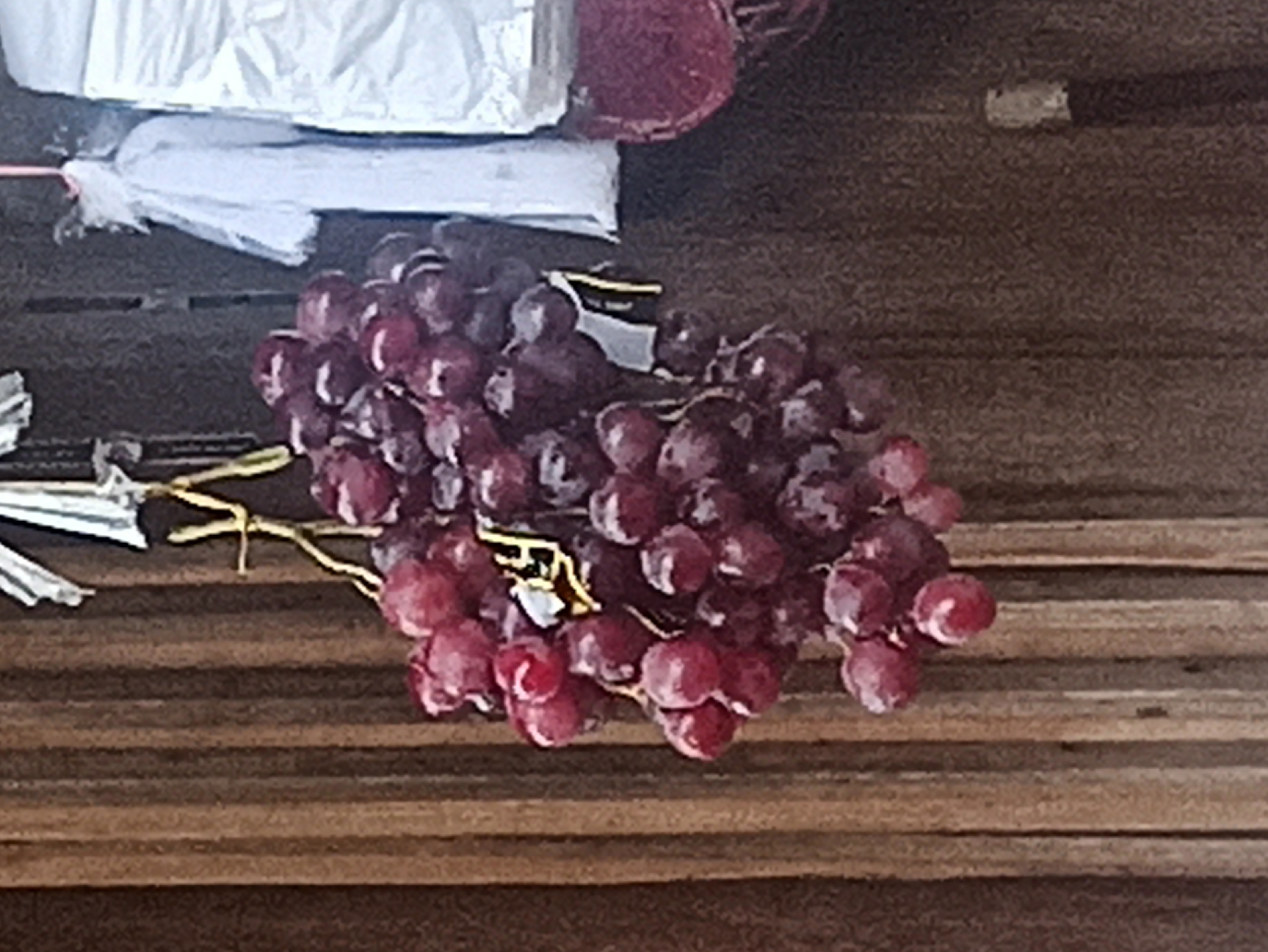A bunch of dark red grapes is hanging from a string, attached to a wooden structure. The grapes are ripe and clustered together. Behind the grapes, a white cloth is draped, and to the right, a red circular object is visible. The overall scene appears to be outdoors, possibly in a market or a roadside stall in the city of Tuba, Indonesia, given the context provided. The wooden structure suggests it might be part of a stall or a building facade.