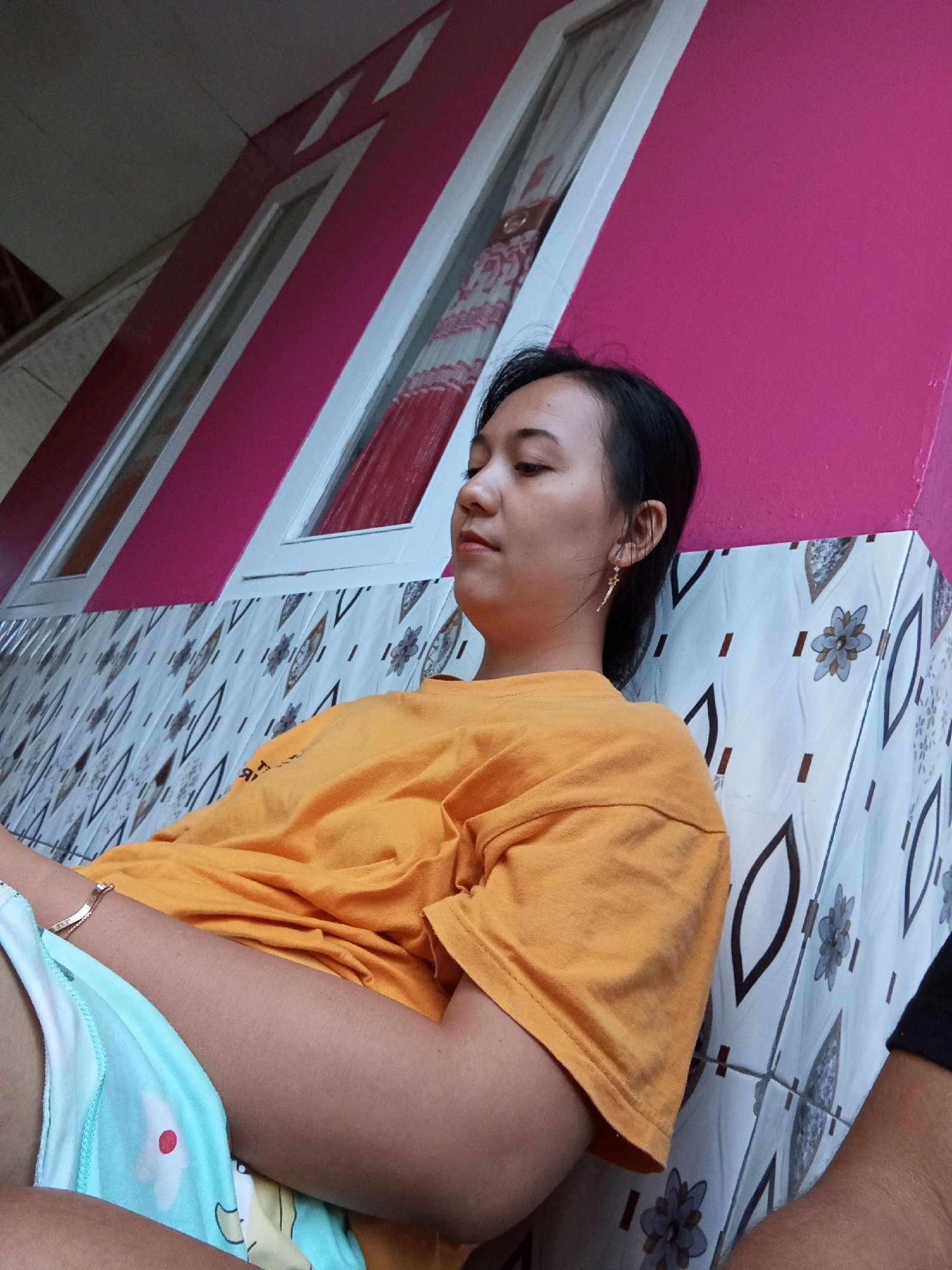 A young woman with dark hair pulled back and wearing a bright orange t-shirt and light blue patterned shorts is sitting with her back against a wall. She has a delicate gold earring in her left ear and a bracelet on her left wrist. The wall behind her is painted a vibrant pink and decorated with what appears to be patterned tiles at its base. The woman's eyes are closed, and her head is tilted slightly downwards, suggesting she is resting or asleep. The overall setting appears to be an outdoor space, possibly a porch or patio, in Muncang, Indonesia. The time of day is indeterminate.