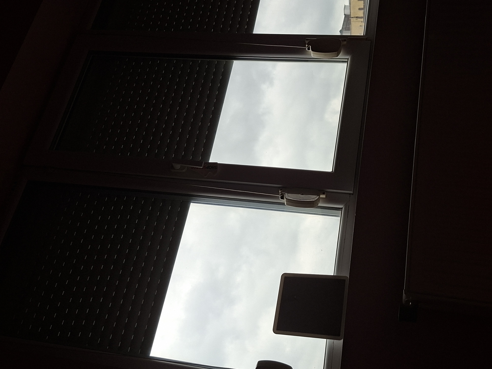 The image depicts a view from inside a building in Pleven, Bulgaria, through a window. The window is partially covered by a closed, dark-colored roller shutter. The uncovered portions of the window reveal a view of a cloudy sky. A rectangular object is attached to the window pane in the lower window. The building's interior is dimly lit.