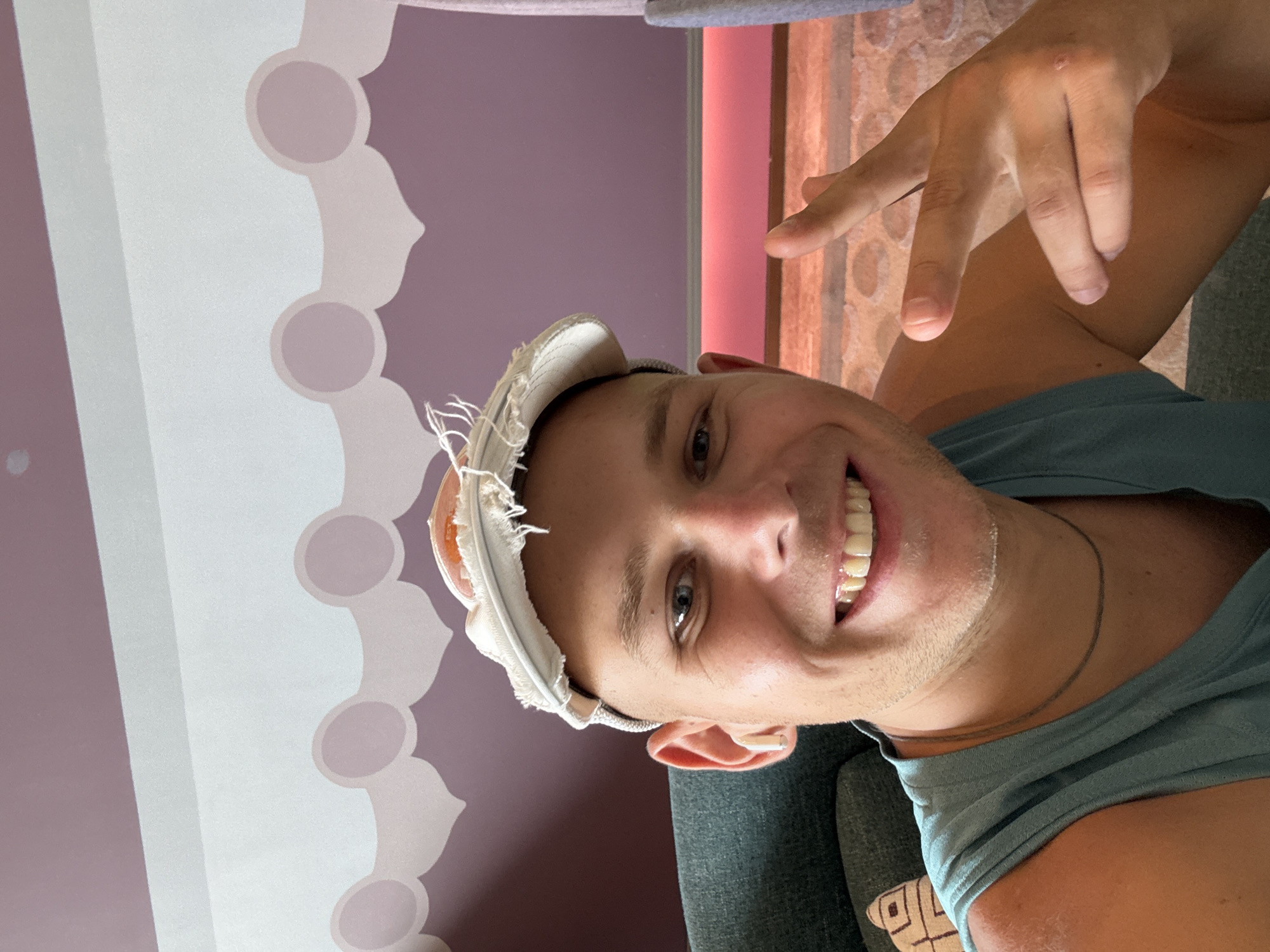 A man with a shaved head smiles for a selfie. He is wearing a blue tank top and a white cap with a fried egg design on it. He has a white wireless earbud in his left ear and is making a peace sign with his right hand. The background is a purple wall with a decorative pattern and a pink light strip. The image was taken in Huangpu District, China.