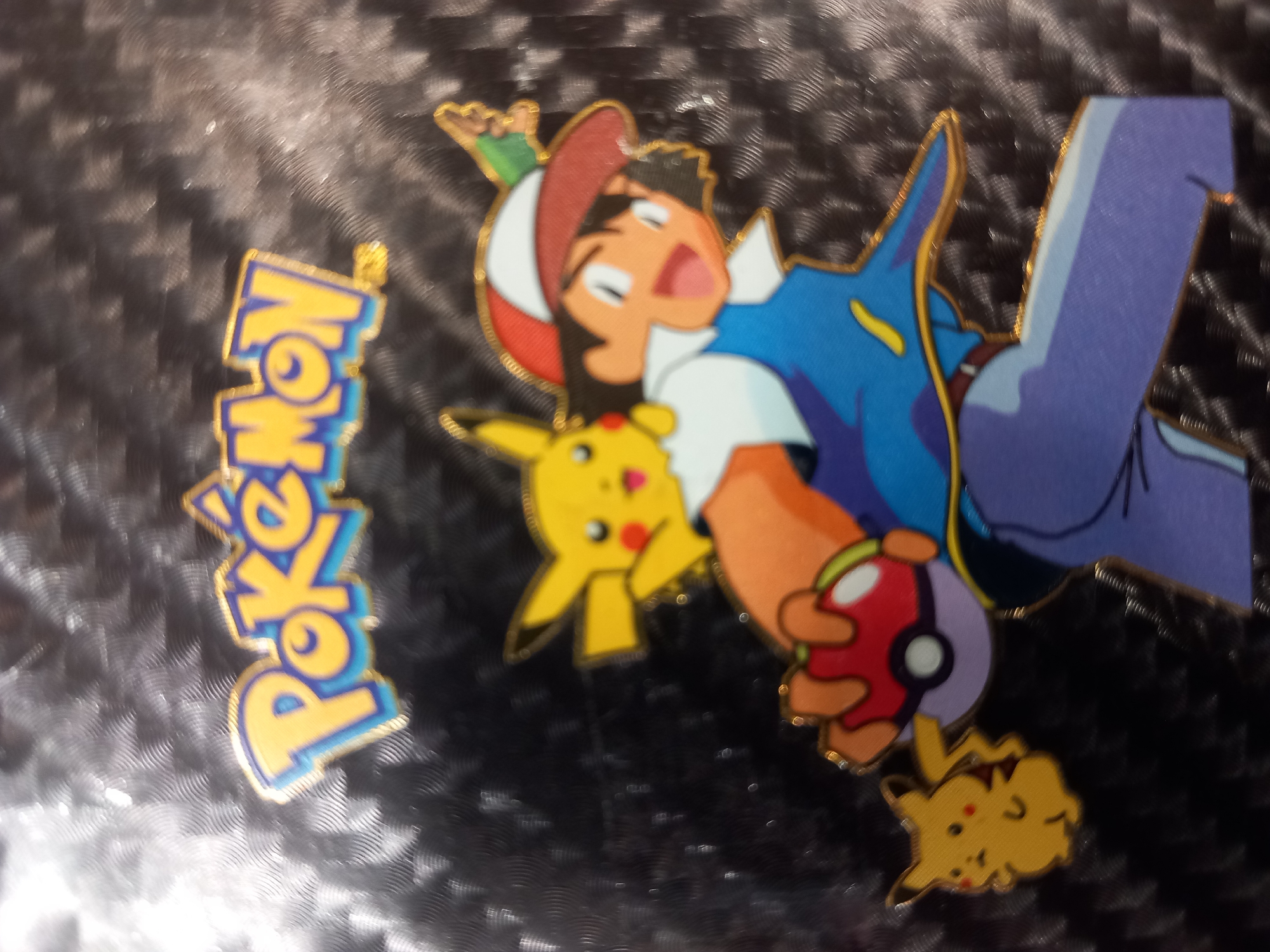 The media file is an image of several Pokemon-themed enamel pins on a dark, textured surface. The primary subject is Ash Ketchum, a character from the Pokemon franchise, depicted in his signature blue vest, red and white hat, and dark hair. He is shown with a wide, excited smile and appears to be in motion, possibly mid-jump or running. Ash is holding a red and purple Pokeball in his right hand.
Next to Ash, and slightly in front of him, is a yellow Pikachu pin, one of Ash's iconic Pokemon companions. Pikachu is also depicted with a happy expression. Below and to the right of Ash, there is another smaller pin of Pikachu, this one appearing to be lounging or sleeping, with its eyes closed and a contented look.
To the left of Ash, vertically oriented, is a pin displaying the word "Pokemon" in stylized yellow letters with blue outlines. The letters are bold and have a metallic gold edge, typical of enamel pins. The overall color palette is bright and vibrant, contrasting with the dark background.
The background has a repetitive, almost ripple-like texture, and appears to be a dark, possibly black or very dark gray material. There are some glints of light on the surface, suggesting it might be slightly reflective or have a metallic sheen.
The image captures a collection of collectible pins, suggesting a fan of the Pokemon franchise. The expressions on the characters' faces and the dynamic pose of Ash convey a sense of adventure and excitement, which are core themes of the Pokemon series. No specific time of day, weather, or location cues are present, other than the fact that these are decorative pins.