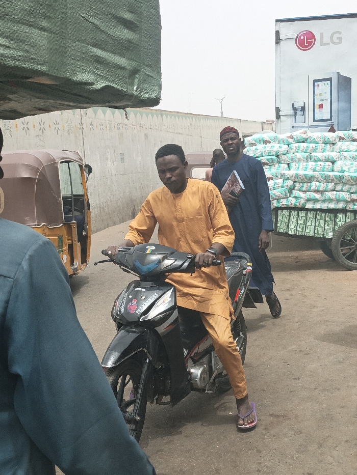 The image displays a daytime street scene in Fagge C, Nigeria. In the central foreground, a male of dark complexion, wearing an orange long-sleeved traditional tunic and matching trousers, is seated on a black and grey underbone motorcycle. He grips the handlebars, and his left foot, wearing a purple flip-flop, rests on the unpaved ground. A dark-colored watch is visible on his left wrist.
To the right and slightly behind the motorcyclist, another dark-complexioned male, dressed in a dark blue traditional garment and a reddish-brown cap, is walking towards the viewer's right. He holds a patterned book or folder in his left hand. The extreme foreground left shows the back and right shoulder of an individual wearing a dark teal long-sleeved garment.
To the left of the central motorcyclist, a brown three-wheeled auto-rickshaw (Keke Napep) is visible. A large object, partially visible in the upper left, is covered with a green tarp. In the background, a light grey concrete wall, adorned with decorative patterns featuring small triangles and star-like shapes, spans the mid-ground. To the far right, a white truck bearing an "LG" logo and a refrigerator graphic is present. Behind this truck, numerous white sacks or bags are stacked high on a two-wheeled cart or trailer. The ground is an unpaved, light brown surface, and the sky is bright and hazy.