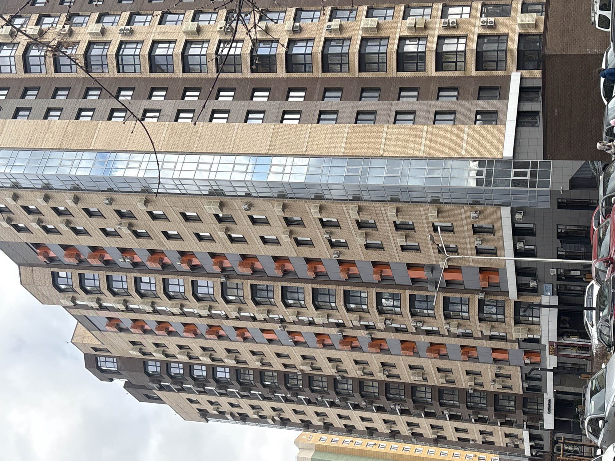 A low-angle shot showcases a multi-story building in Krasnoyarsk, Russia. The building features a varied architectural design, with alternating tiers of light-brown brick and darker brown brick. Some windows are flanked by air conditioning units, while others are framed by orange panels. A horizontal band of glass windows, likely for an office space, cuts across the mid-section of the building. The right edge of the frame captures a few parked cars, while a street lamp rises along the same edge. A branch of a tree is visible at the top-left of the frame. Overcast skies are seen above.