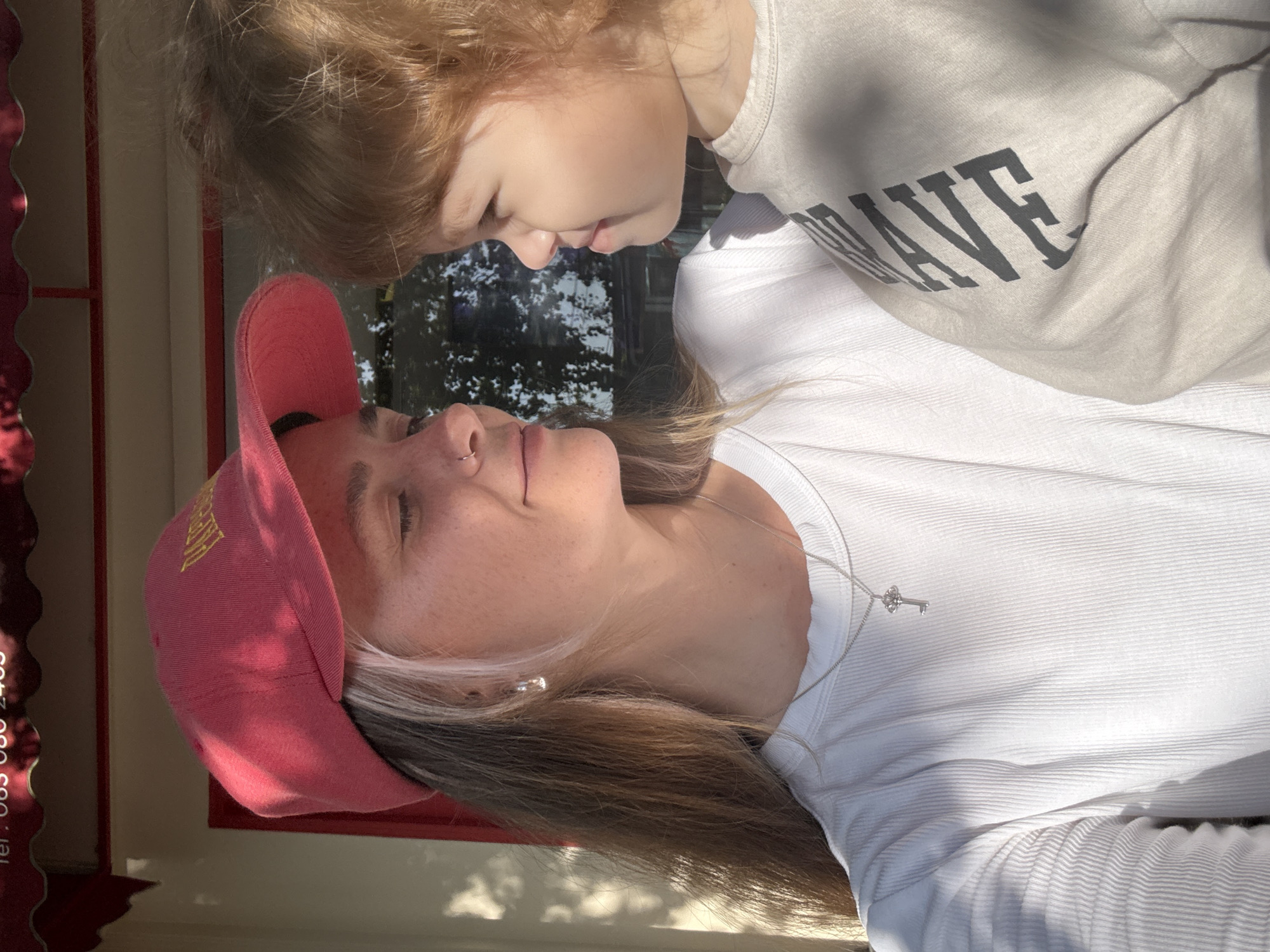 A close-up, medium shot captures two individuals, a woman and a young child, outdoors in what appears to be Choeng Thale, Thailand. The woman, wearing a red baseball cap and a white ribbed long-sleeved shirt, has her hair down and is looking at the child. She has a nose ring and a silver necklace with a key pendant. The child, with light brown hair, is wearing a light-colored long-sleeved shirt with dark text on the sleeve that reads "WAVE". They are positioned in profile, facing each other and appear to be interacting affectionately. The background consists of blurred foliage and a building with red trim, suggesting an outdoor setting possibly near a cafe or shop. Sunlight casts shadows on the subjects and the environment, indicating it's daytime.