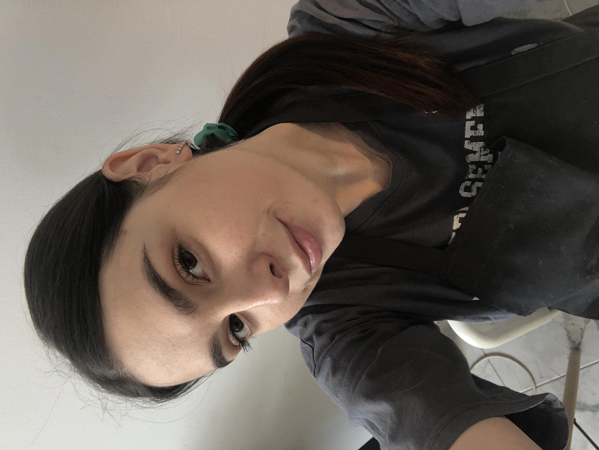 A young woman with long dark hair pulled back in a green scrunchie is pictured in a close-up, slightly tilted selfie. She wears a gray t-shirt with white lettering, partially obscured. Her skin is fair, and she has dark eyebrows and brown eyes. Her ears are visible, with an earring in the upper ear cartilage. The background includes a white wall and a glimpse of a white chair and flooring. There is no direct evidence indicating this photograph was taken in Chaikovsky, Russia.
