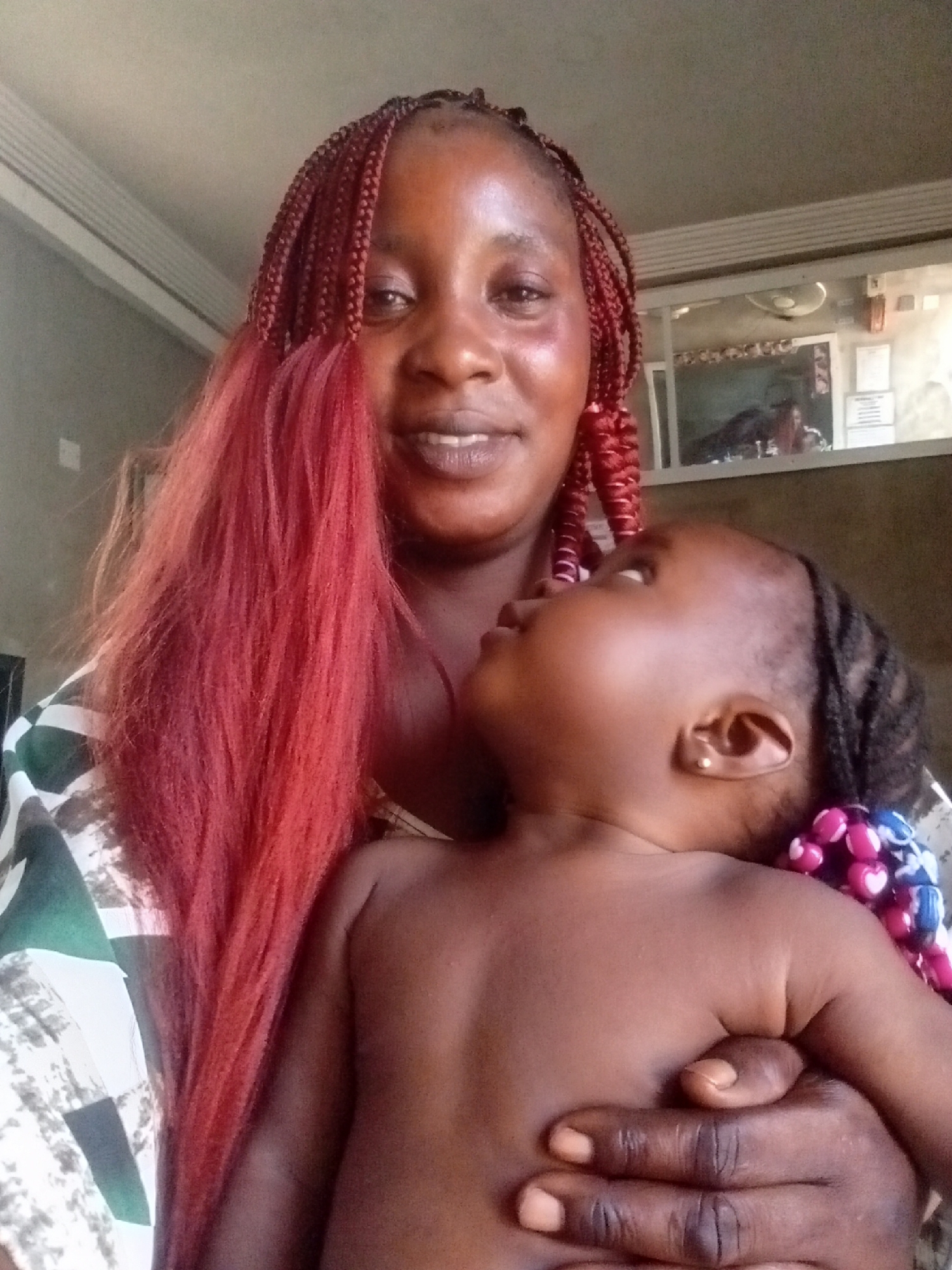 This image captures a woman holding a young child indoors, likely in Akwanga, Nigeria. The woman is positioned centrally, looking towards the camera with a gentle, slight smile. She has distinctive red braided hair, with some sections of the braids appearing as loose, flowing red strands on her left side. She wears a garment with a bold pattern featuring green, white, and black geometric shapes.
The child, appearing to be a toddler or baby, is held against the woman's chest, looking upwards with a curious expression. The child is bare-chested and has small, dark braids adorned with colorful beads in shades of pink, purple, and blue. A small gold stud earring is visible in the child's left ear. One of the woman's hands is wrapped around the child, providing support.
The setting appears to be an indoor space, possibly a home or a commercial establishment. A plain, light-colored ceiling is visible above. In the background, there is a window or display case with a reflective surface, revealing some indistinct objects like a possible fan or light fixture, and some papers or signs on a wall behind it. No specific text is legible on these background elements. The overall impression is one of affection and warmth, captured during what appears to be daytime.