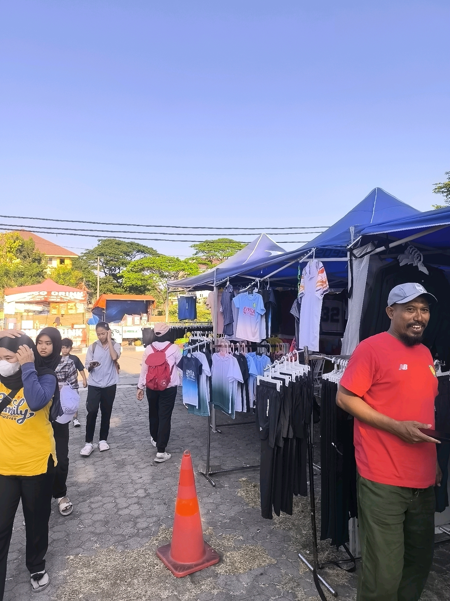 The image depicts a street market scene in Surabaya, Indonesia, under a clear blue sky. In the foreground, a man with a mustache, wearing a red t-shirt, a grey cap, and green pants, smiles at the camera. He is standing next to a market stall displaying various clothing items, including t-shirts and shorts, hanging on racks under a blue tent.
Several people are walking past the stall. To the left, a group of women are seen. One woman in the front wears a yellow t-shirt and black pants, with a blue jacket draped over her shoulder. Beside her, another woman wears a black hijab and a black top. Further back, more individuals, including a young boy in a checkered shirt, are walking away from the camera.
In the background, there are other market stalls, including one with the text "GO DRINK" visible. Buildings and lush green trees are also present, suggesting an outdoor urban environment. A bright orange traffic cone is placed on the cobblestone ground in the middle of the scene. The lighting indicates it might be late afternoon.
