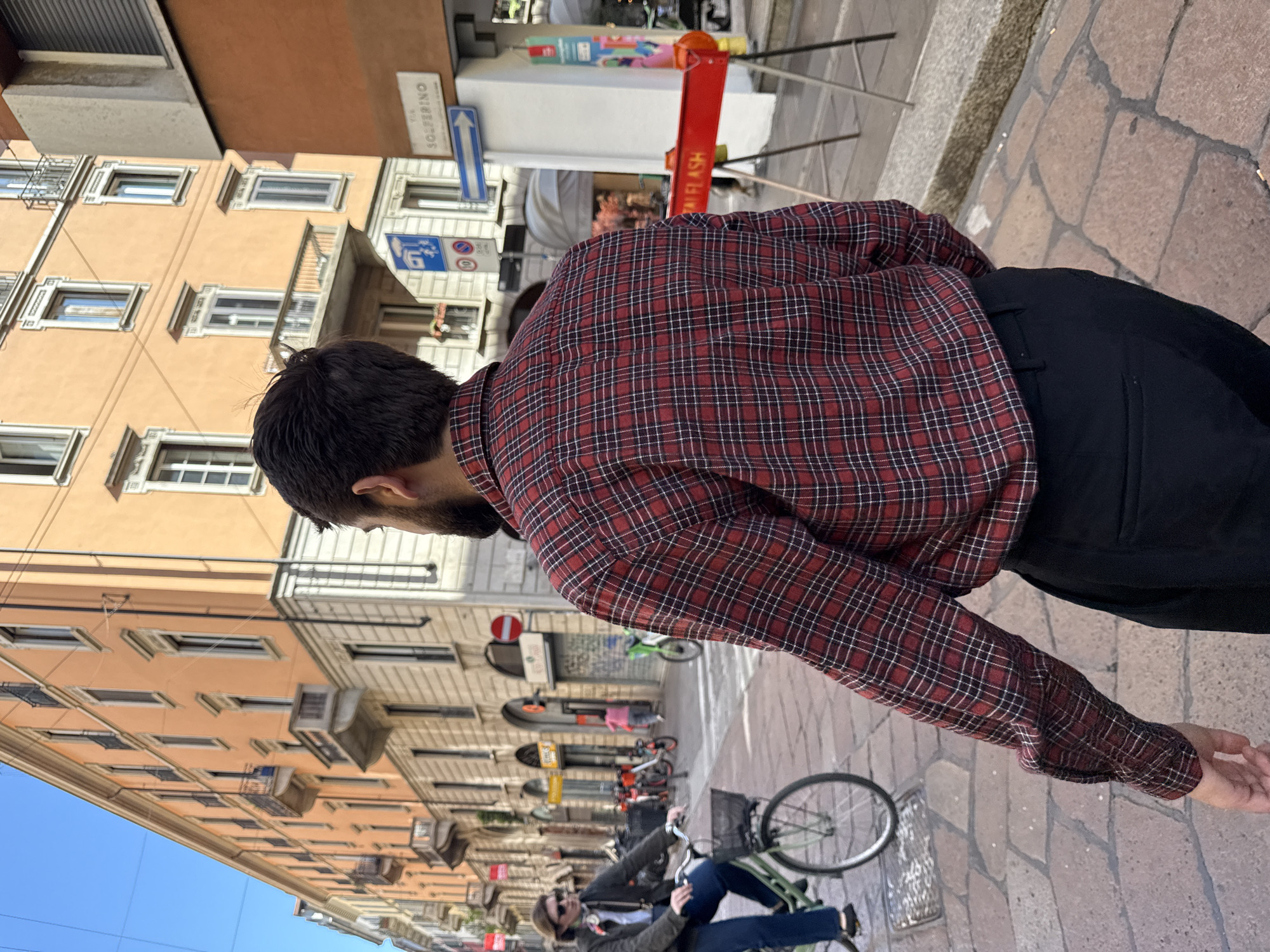 The image shows a man in a red and black plaid shirt and black pants standing on a cobblestone street. He has dark hair and a beard and is facing away from the camera, looking down the street. In the background, there is a street scene with buildings, bicycles, and other people. A street sign for "Solferino" is visible, suggesting the location is in the city of Milan, Italy. There is a sign that says, "ITAL FLASH". One person is riding a green bicycle. The sky is blue.