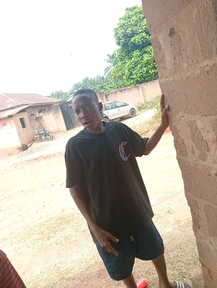The scene is set in a semi-urban or rural residential area within Eziudo, Nigeria. A young individual, appearing male, with short dark hair and dark skin, stands in the immediate foreground, slightly angled to the left, with their head turned towards the viewer. They are wearing a black short-sleeved t-shirt with a red and blue outlined "C" logo on the chest, dark blue shorts, and a black and white striped slide sandal on their visible left foot. The individual's right hand rests on a rough-textured, light brown concrete pillar or wall that occupies the right side of the frame. A red plastic cup is on the ground near their left foot.
The ground is unpaved, consisting of light brown dirt and sparse dry vegetation. In the mid-ground, a light-colored (possibly silver) minivan or SUV is parked. Behind it, a low, light brown concrete wall extends horizontally. Further left, a single-story building with a light brown facade and a darker roof is visible, along with a closed dark metal gate and a light blue or grey motorcycle parked nearby. Dense green foliage and tall trees, including a prominent leafy tree, are present in the background, under a bright, likely overcast sky.