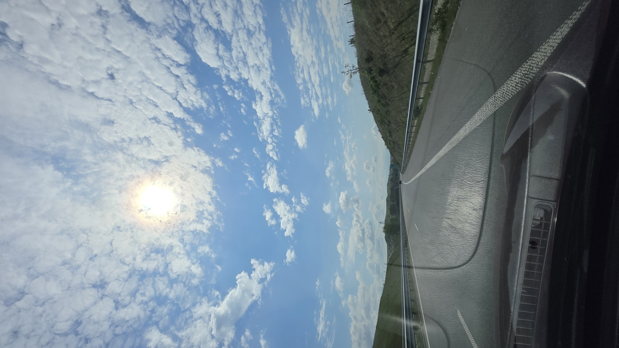 The image presents a view from inside a vehicle, possibly on a highway in Leshnitsa, Bulgaria. The road stretches out to the right, marked with white lines, next to a metal guardrail and grassy terrain. The sky occupies the left side of the frame, with scattered white clouds and a bright sun. Part of the vehicle's dashboard is visible in the lower right corner. There are no people immediately visible in the frame.