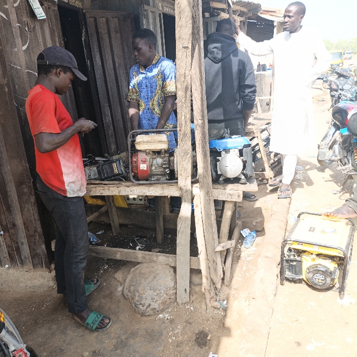 The image depicts an outdoor scene in Sabon Gari East, Nigeria, characteristic of a street-side repair or sales area. Four adult males are visible under daylight conditions.
In the left foreground, a male wearing a black baseball cap, a red short-sleeved t-shirt with a white stain on the front, dark trousers, and green "GUCCI" slides, stands leaning slightly forward, appearing to examine a small, dark object held in both hands. Directly behind him, positioned behind a rustic wooden workbench, a male in a blue and yellow patterned short-sleeved shirt stands, observing the first individual.
On the wooden workbench, two portable generators are present: a red and beige unit on the left and a blue and white unit on the right. Directly beneath the workbench, a large, dark, irregularly shaped stone object rests on the dusty ground, surrounded by scattered debris, including a discarded plastic bottle.
Further to the right, a male in a black hooded sweatshirt stands with his back partially towards the viewer, also positioned behind the workbench. To his immediate right, a fourth male, dressed in a white long-sleeved tunic and light-colored sandals, stands facing right, with his right arm raised and hand resting on an unseen surface or gesturing.
In the right foreground, a yellow and black portable generator with a metal frame rests on the dirt ground. The background reveals additional street or market activity, including several motorcycles and rudimentary structures made of corrugated metal and wood. The ground throughout the scene is visibly dusty.