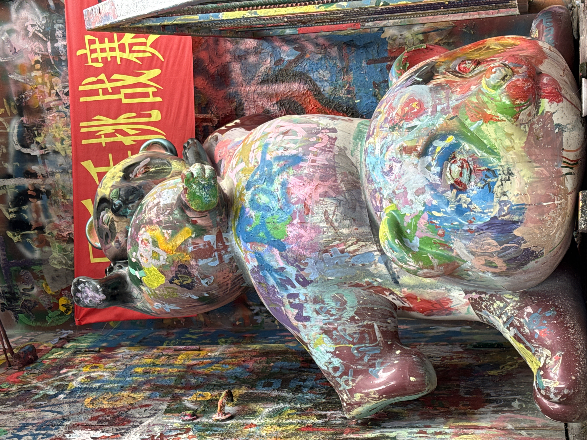 The image depicts two large, heavily paint-splattered teddy bear sculptures positioned within an intensely painted environment in Jiulongpo District, China. The upper sculpture is partially visible, showing its multi-colored, paint-dripped body and head, with one dark, paint-marked limb extending upwards. The lower sculpture occupies the foreground, presenting its head and upper body, entirely covered in thick, layered applications of various paint colors including red, blue, green, yellow, pink, and white. Its underlying form on visible limbs is a deep reddish-brown. The surrounding walls are completely coated in dense, textured layers of dried, multi-colored paint, exhibiting extensive drips and abstract patterns. A red banner with yellow Chinese characters, "千挑战赛" (Thousand Challenge Competition), is stretched horizontally behind the upper sculpture. A metallic structure, partially painted, is visible on the far right. A small, light-colored dish or object rests on the ground in front of the lower sculpture's head. Small, paint-covered protrusions are present on the left wall.