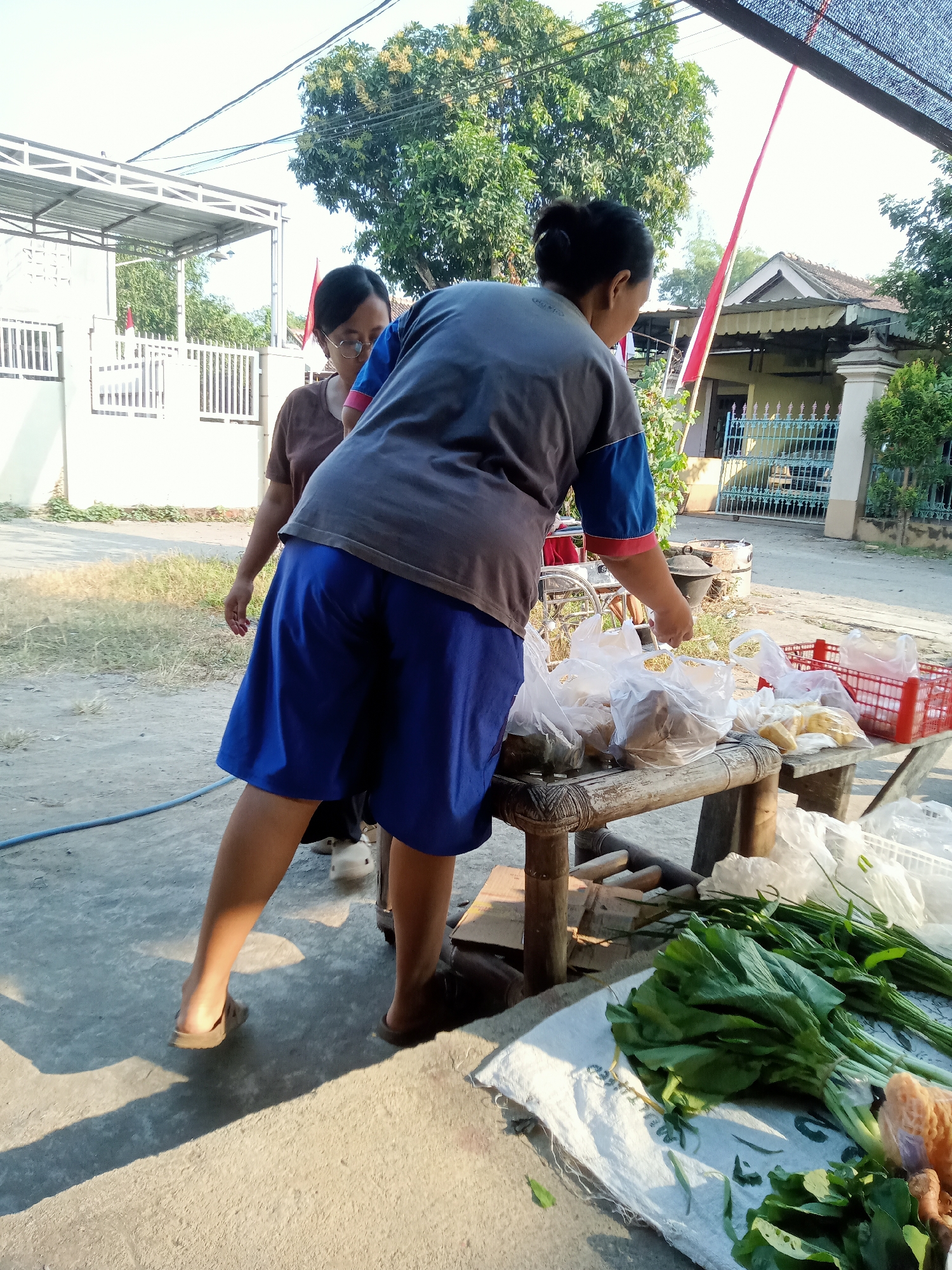 In Simo, Indonesia, two women are standing at a makeshift market stall in what appears to be an outdoor residential area. The woman in the foreground, wearing a grey t-shirt with blue and red trim and blue shorts, is reaching for items on the stall. The stall is made of bamboo and wood, covered with a woven mat, and displays various goods in plastic bags, including produce like leafy greens and what looks like ginger or turmeric. A red plastic crate is also visible. Behind her, another woman with glasses, wearing a brown t-shirt, looks towards the stall. In the background, residential buildings with white fences and a car are visible, along with trees and electrical wires. The ground is concrete and dirt, with shadows cast by the bright sunlight. A blue hose lies on the ground.