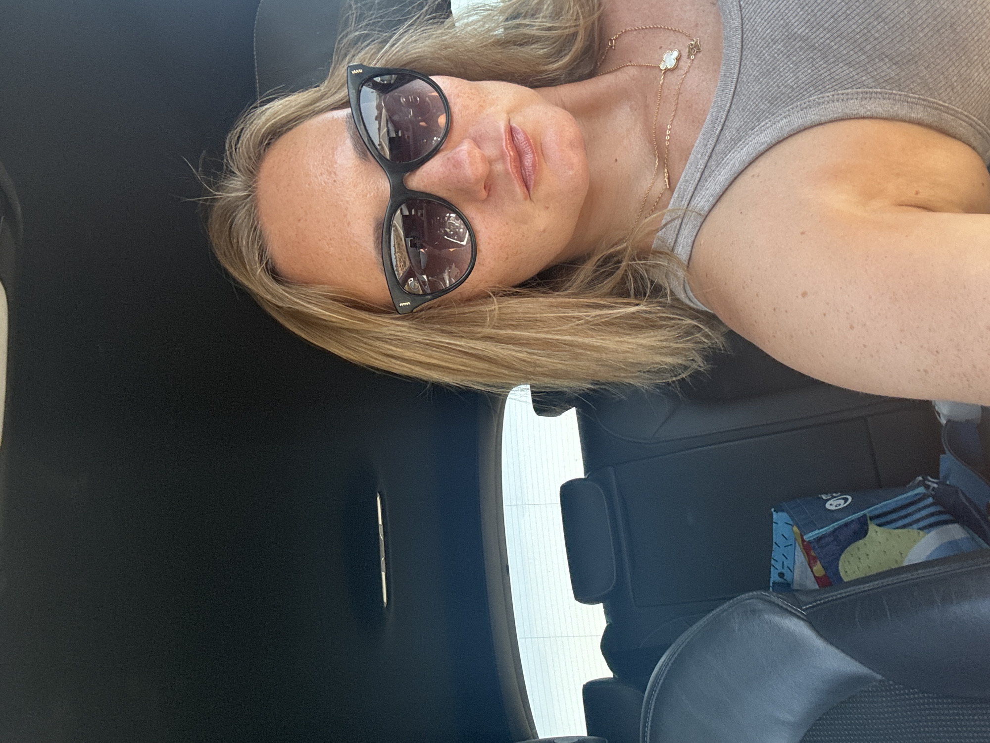 The image shows a woman in the passenger seat of a car. She is wearing sunglasses and a grey tank top. Her blonde hair is loose around her shoulders. She has freckles on her face and arms. A colorful tote bag is visible on the seat next to her. The background shows the interior of the car, including the headrests of the back seats and the car's headliner. The woman appears to be taking a selfie. The location is identified as Olhão, Portugal.