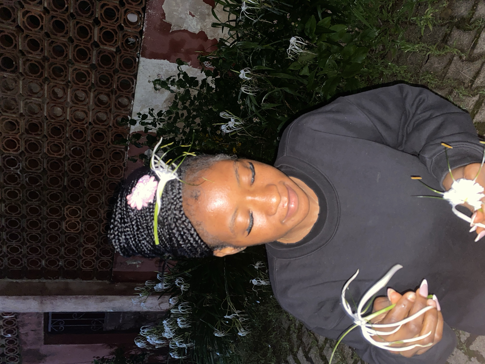 A young Black woman with braided hair adorned with a pink flower and a green ribbon is in a dimly lit outdoor setting, likely at night. She is wearing a black sweatshirt and holding white spider lilies in her hands. She is looking down with a slight smile. In the background, there is a textured wall with circular patterns, a doorway, and lush green foliage with more white flowers. The ground appears to be paved with bricks. The overall ambiance suggests a tranquil moment in a garden or courtyard, possibly in Jos, Nigeria, given the specific architectural style of the wall which is common in some parts of the country.