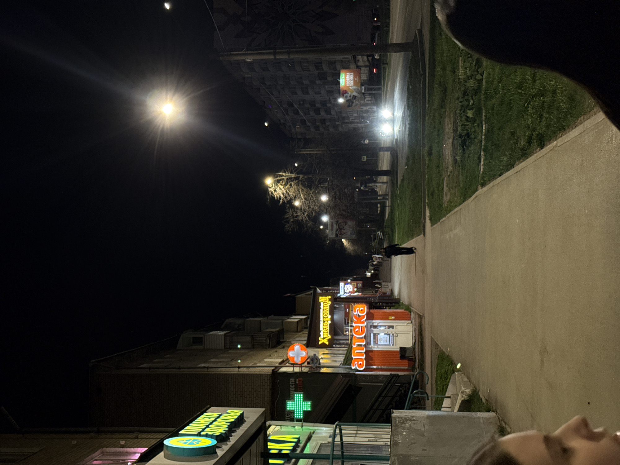 This nighttime image shows a street scene in Perm, Russia. A sidewalk runs along the right side of the frame, bordered by a strip of grass. On the left side of the sidewalk, there are several storefronts with brightly lit signs in Russian. A person is walking on the sidewalk in the distance. Streetlights illuminate the scene. There is a building visible in the background with a mural on its side. The sky is dark with a bright light source, possibly a streetlamp or the moon. In the foreground, part of a person's face is visible in the lower right corner.