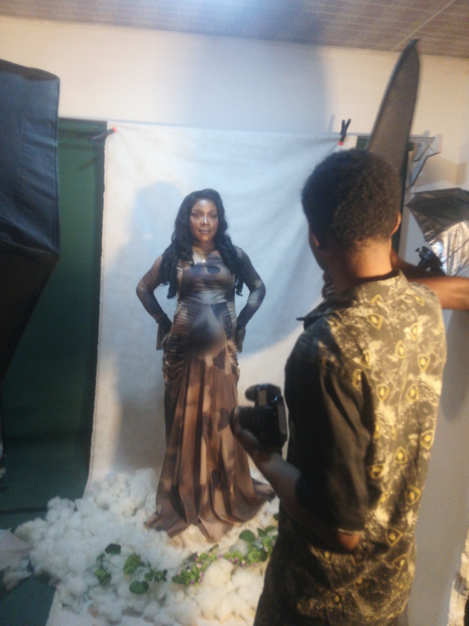 In an indoor photography studio, a photographer is capturing a pregnant woman posing for a picture. The woman is standing in the center of the frame, wearing a flowing, earth-toned maternity dress. She has long, dark hair and is looking towards the camera. In front of her, the floor is covered with what appears to be cotton batting, simulating clouds, with some green foliage and small purple flowers scattered around. To the left of the woman, a large, black softbox or light modifier is visible. To the right of the frame, the photographer, seen from the back, is holding a camera and appears to be in the process of taking a photograph. The background consists of a plain white sheet, possibly a backdrop.