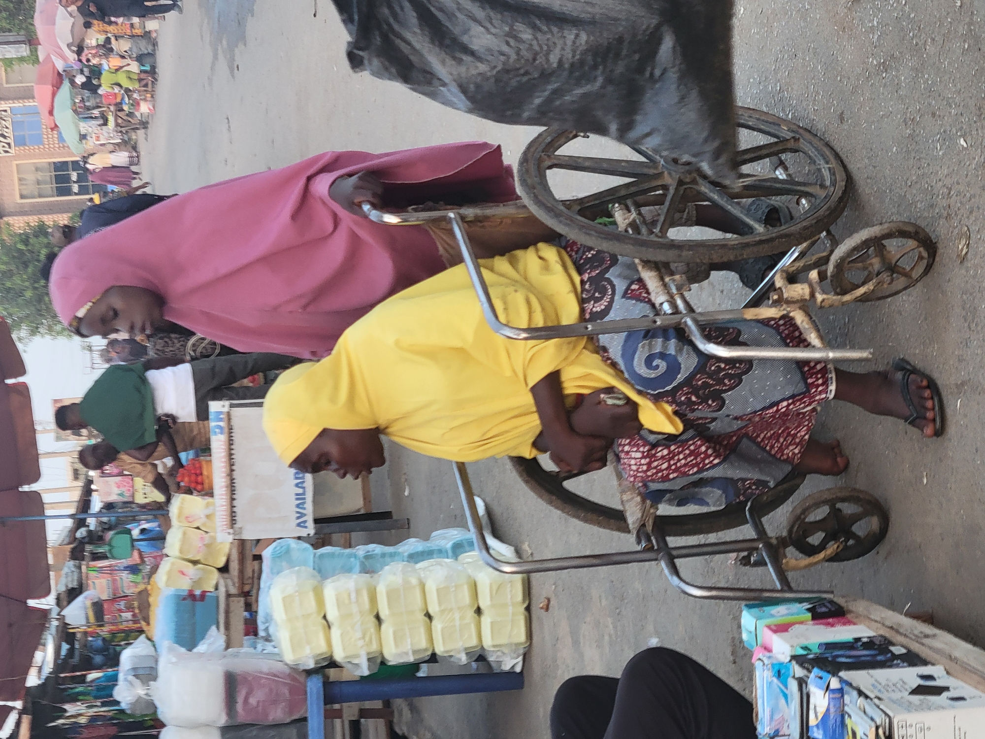 Here's a detailed description of the image:
**Setting:** An outdoor market scene in Madalla, Nigeria.
**Subjects:** Two African women are the primary focus, one wearing a yellow headscarf and the other a pink one. Both are sitting on an old, metal-framed cart with wheels. A third person, wearing black pants, is partially visible in the foreground. There are several other people visible in the background, further back in the market.
**Objects:** The cart is loaded with various items including rectangular packages wrapped in clear plastic. Additional merchandise, including packaged food items, is arranged on a shelf next to the cart. The street and buildings form the background.
**Actions and Interactions:** The woman in the yellow headscarf is sitting on the cart and appears to be holding her knees. The woman in the pink headscarf is behind her. The cart appears to be part of their market setup. The other people in the background are likely shoppers or vendors.
**Environment:** The location is a busy marketplace with various vendors and shoppers. The ground appears to be paved. The lighting suggests it is daytime.