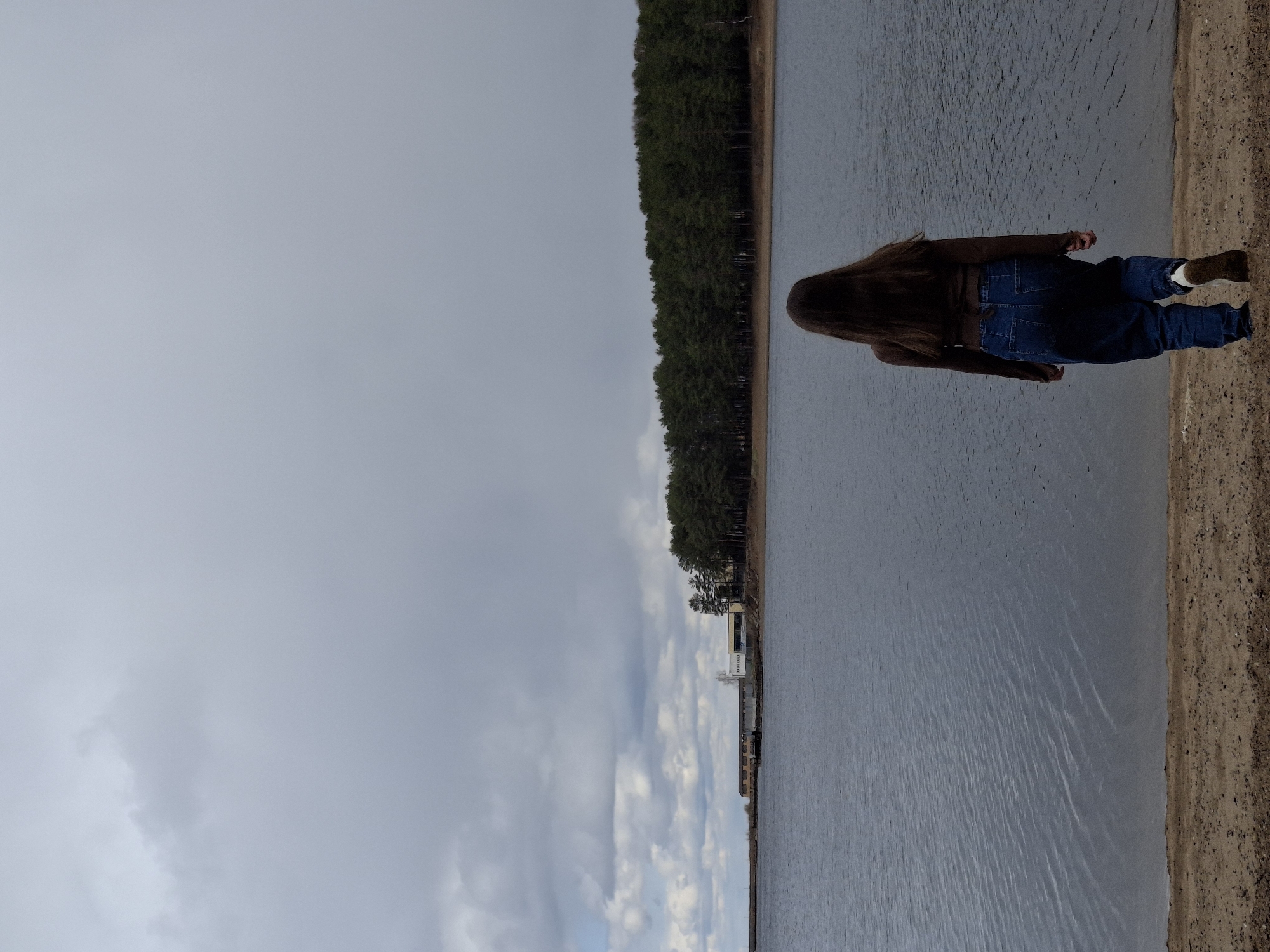 The image displays an outdoor scene featuring a solitary person on the bank of a large body of water, under an overcast sky. The photograph is taken from behind the person, who is looking out across the water towards the distant shore.
The subject is a person with long, dark brown hair, wearing a dark brown jacket, blue jeans, and brown boots with visible white cuffs. They stand on a sandy or muddy bank that lines the water's edge in the foreground and along the right side of the frame.
The middle ground is dominated by a vast body of water, likely a reservoir or large river, which exhibits subtle ripples on its surface, reflecting the muted light from the sky. In the background, a dense line of dark green coniferous trees stretches horizontally across the distant shore. Beyond or amidst this treeline, several structures are visible, including a prominent white building. Closer inspection of the building reveals partial Cyrillic text, reading "НЕФТ...", suggesting an industrial presence such as an oil facility (НЕФТЕБАЗА - oil depot).
The sky above is uniformly grey and overcast, indicating a cloudy daytime. Despite the lack of direct sunlight, there's a soft, diffused light over the scene. The overall mood is quiet and contemplative, with the person seemingly observing the expansive landscape. The location context of Chaikovsky, Russia, aligns with the depiction of a large reservoir and an industrial shoreline.