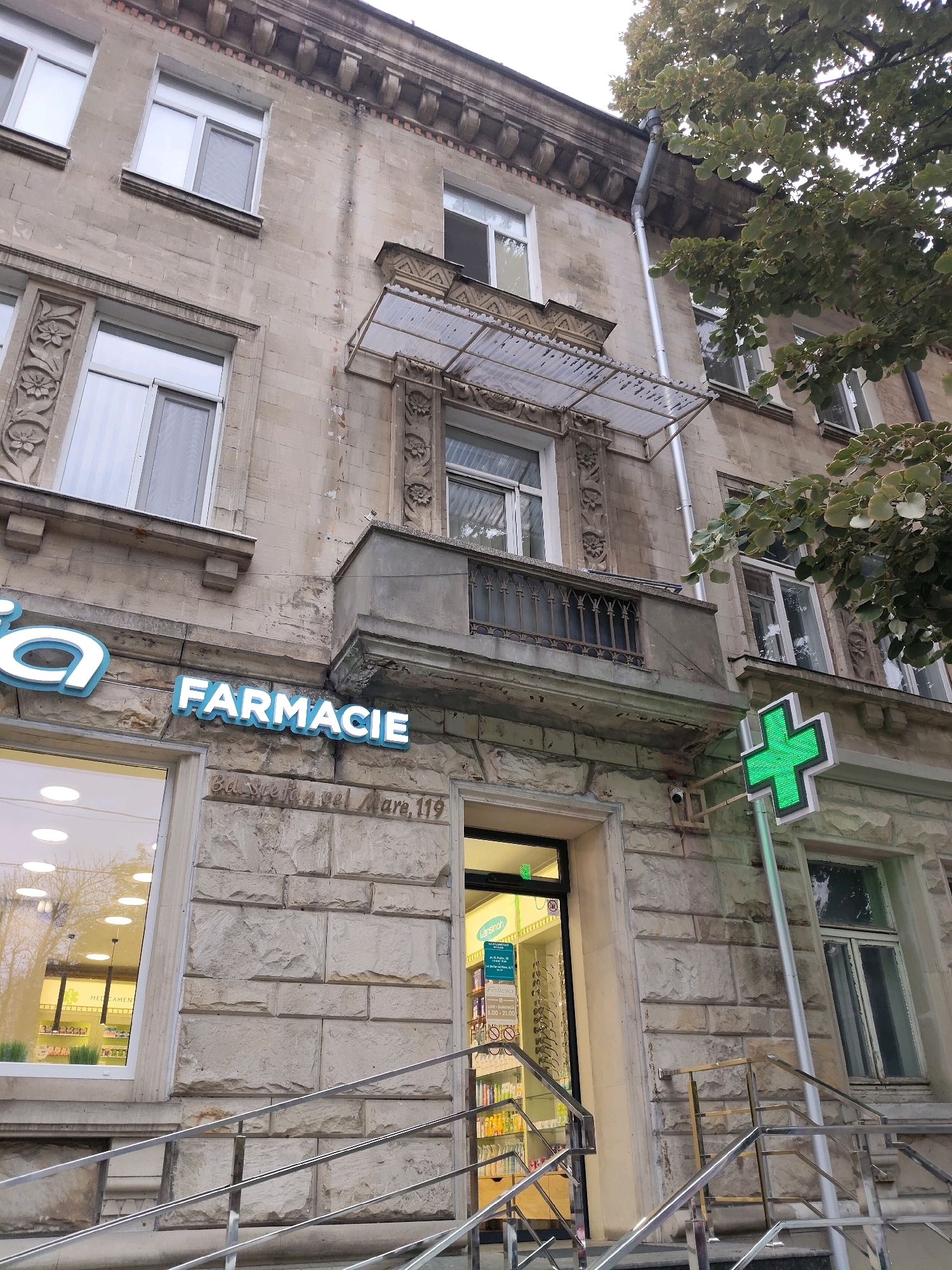 A low-angle, eye-level exterior shot shows the facade of a building with a pharmacy. The building appears to be made of stone blocks and has multiple windows. There is a visible sign for "FARMACIE" in blue illuminated letters. A prominent green cross sign, also illuminated, is mounted on a pole outside the building. The entrance to the pharmacy is visible, with shelves of products displayed inside. A set of metal railings leads up to the entrance. Trees with green foliage are visible on the right side of the frame, partially obscuring the upper portion of the building. The image depicts a commercial establishment in the city of Durlești, Moldova.