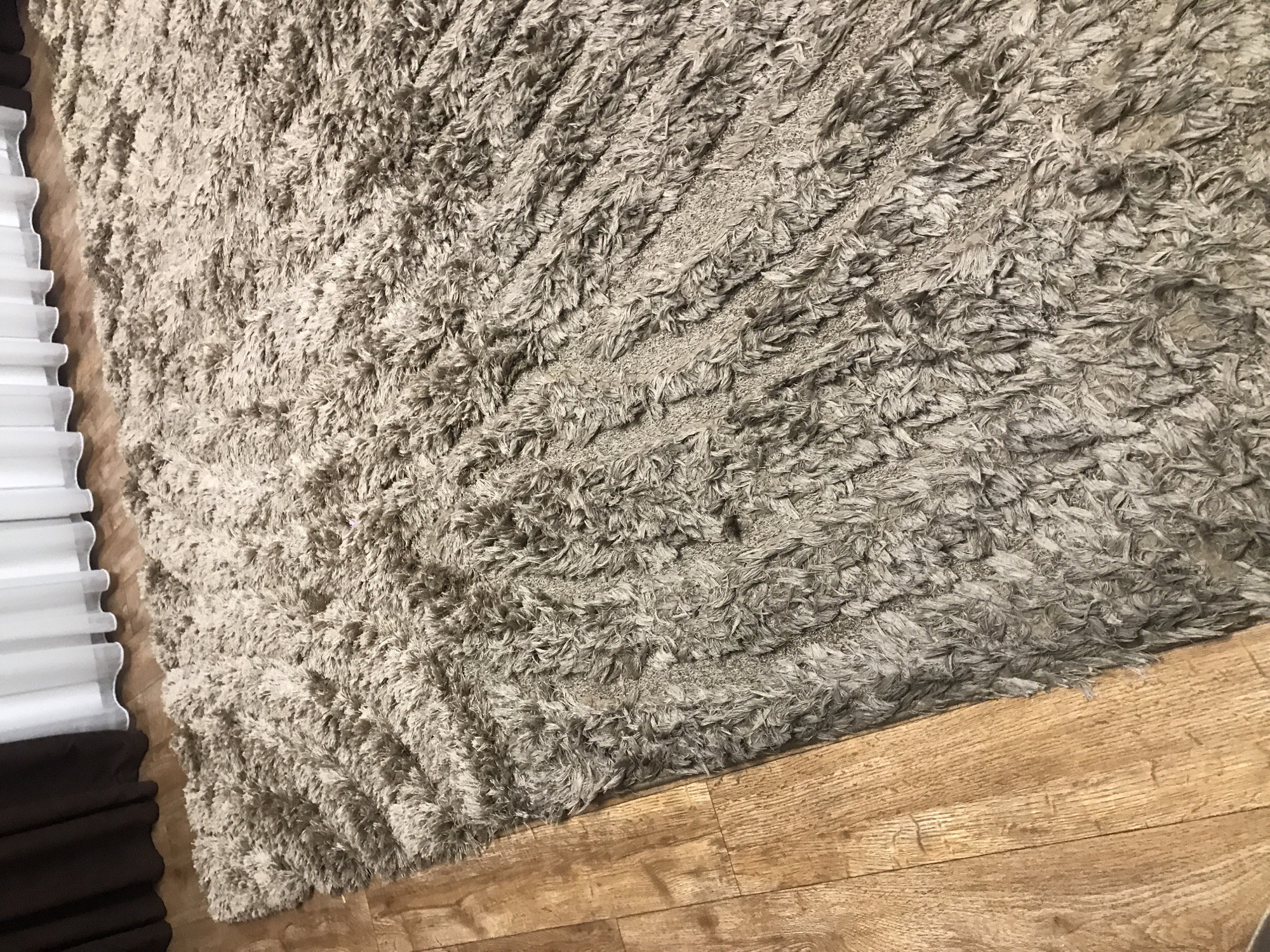 The image shows a portion of a room with a light-colored, shaggy rug partially covering a light brown, wood-grain floor. A white and dark brown window covering is visible along the left edge of the frame. The scene appears to be indoors. No people are visible. There is no location information from Yerevan, Armenia.