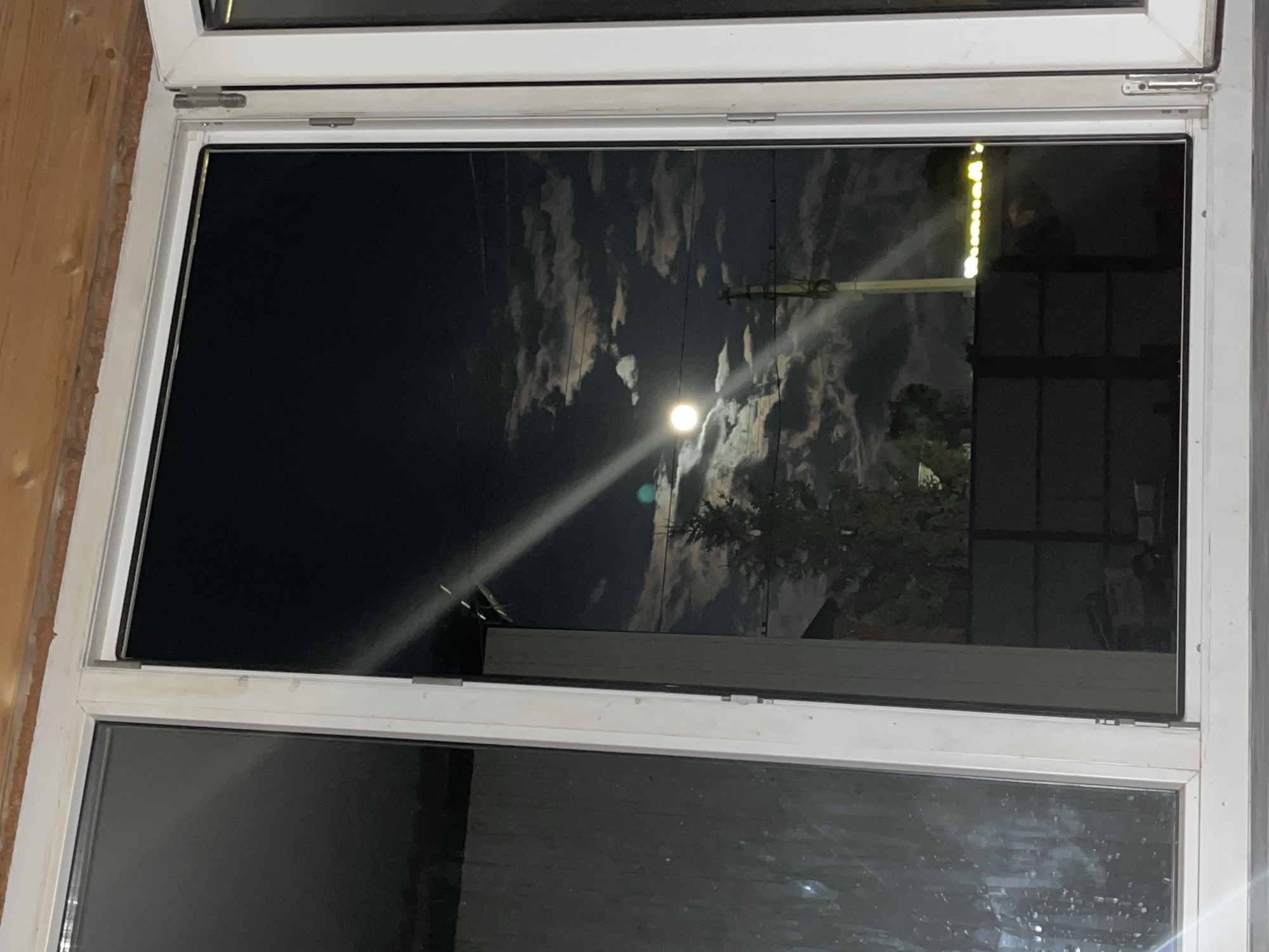 The image shows a night view through a window, possibly in a residential building in Saratov, Russia, given the typical architecture and lighting. The primary focus is the sky, which is dark with scattered clouds illuminated by a bright full moon. A distinct beam of light, likely from a streetlamp or another artificial source, cuts across the moon and clouds from the top left to the bottom right.
Below the sky, silhouetted against the faint moonlight, is a tall, evergreen tree, possibly a fir or pine. Beyond the tree, a dark fence is visible, with the indistinct shapes of buildings and possibly a utility pole with visible wires in the background. Reflections on the window pane obscure some details of the exterior scene, creating a layered effect. The window frame itself is white and appears to be made of uPVC, typical of modern installations. The overall impression is a serene, moonlit night with a touch of urban ambiance.