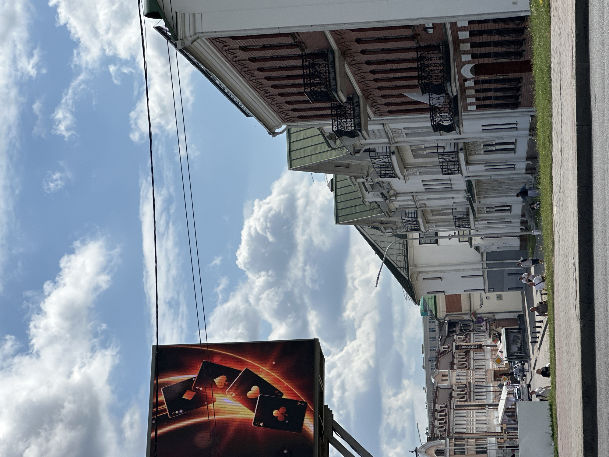 In Minsk, Belarus, a billboard featuring playing cards is visible against a sky with scattered clouds. Adjacent to the billboard, a row of white buildings with green roofs and ornate balconies lines a street. Further down the street, more buildings of varying architectural styles can be seen, including some with brick facades. Several people are walking or standing on the sidewalk, and cars are present on the street. The scene suggests an urban environment with a mix of modern and older architecture.