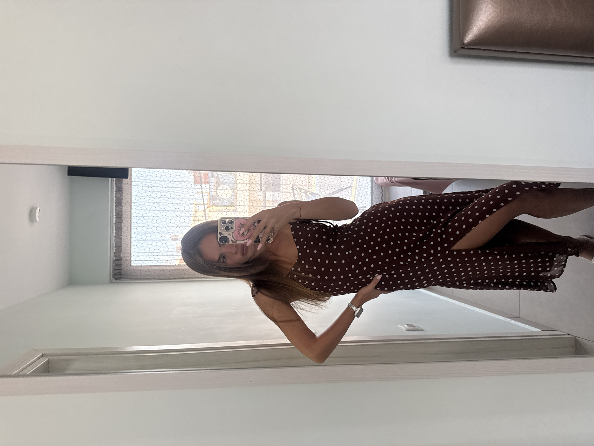 A young woman with long brown hair, wearing a dark brown polka dot midi dress with a thigh-high slit and thin straps, stands in a hallway and takes a selfie in a mirror. She has her right hand on her hip and is wearing a watch on her left wrist. The hallway has light blue walls and a white door frame. Through a window or doorway behind her, a glimpse of a building with arches is visible. The floor appears to be tiled.