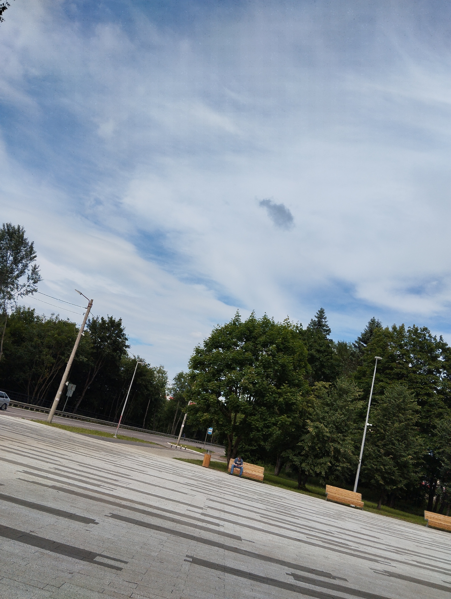 A tilted, full shot captures an outdoor scene with a paved area in the foreground and a line of trees and a road in the mid-ground, under a partly cloudy blue sky. The paved area is made of large, light-grey rectangular tiles, with darker grey rectangular patterns forming lines that recede into the distance. Several wooden benches with backrests are placed along the edge of the paved area, near the trees. One bench has a single person sitting on it, looking down. In the mid-ground, a road runs horizontally, bordered by a metal fence and grass. Beyond the road, a dense line of green trees forms a backdrop, with some taller trees visible in the distance. A lamppost stands on the left side of the paved area, near the road. The sky is a pale blue with wispy white clouds and a darker, small cloud formation towards the top center. The image has a slightly grainy texture. This scene is located in Kingisepp, Russia.