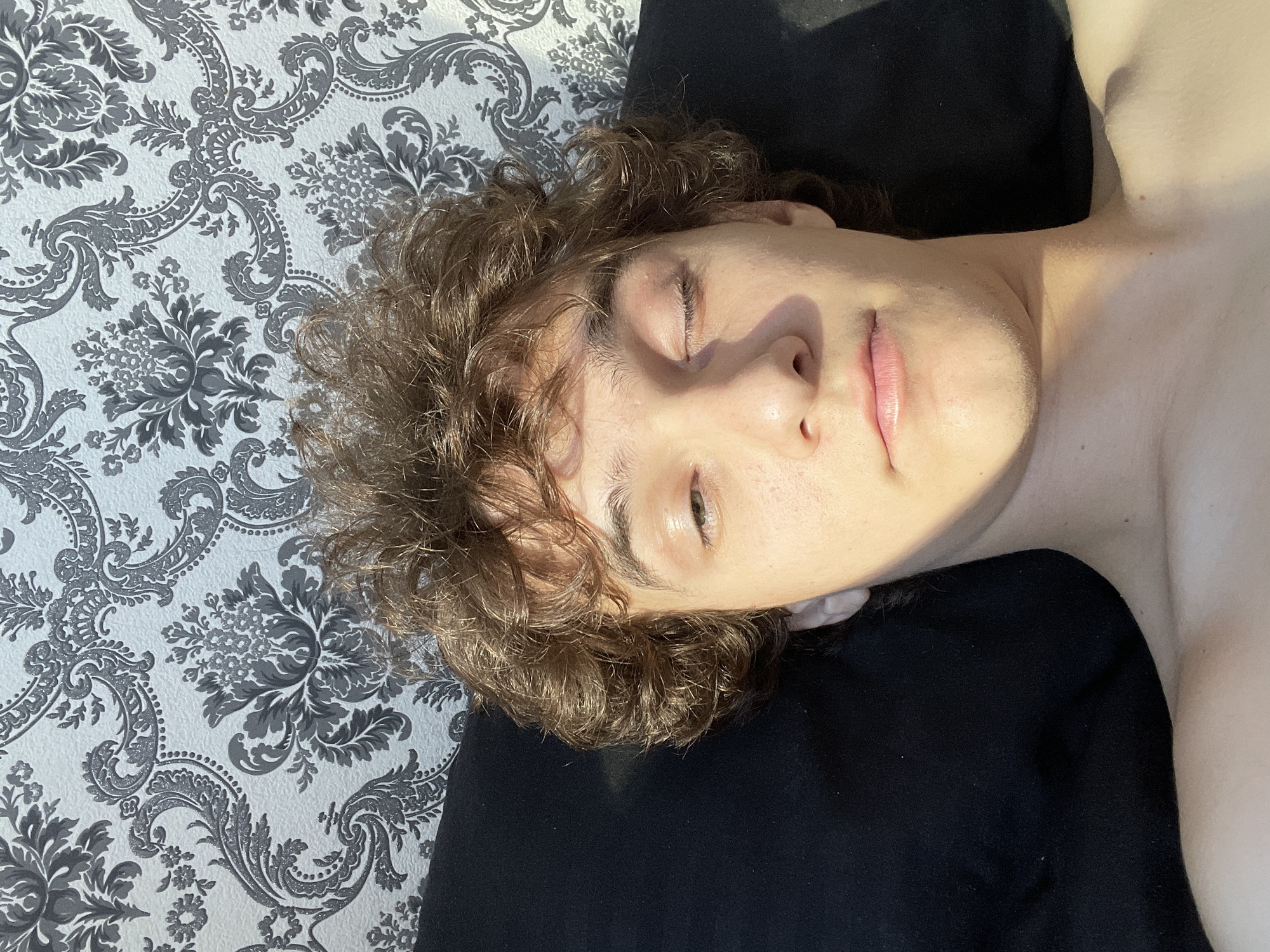 The image is a close-up, overhead, selfie-style photograph of a young person lying on a black surface, possibly a pillow or bed, against a patterned wallpaper. The person has curly brown hair and is looking towards the camera with one eye partially closed. Sunlight casts a diagonal shadow across their face. The wallpaper behind them features a repeating, ornate floral damask pattern in shades of gray and white. The person appears to be indoors, in a room with natural light. There is no visible indication of the location being in Krasnoyarsk, Russia.