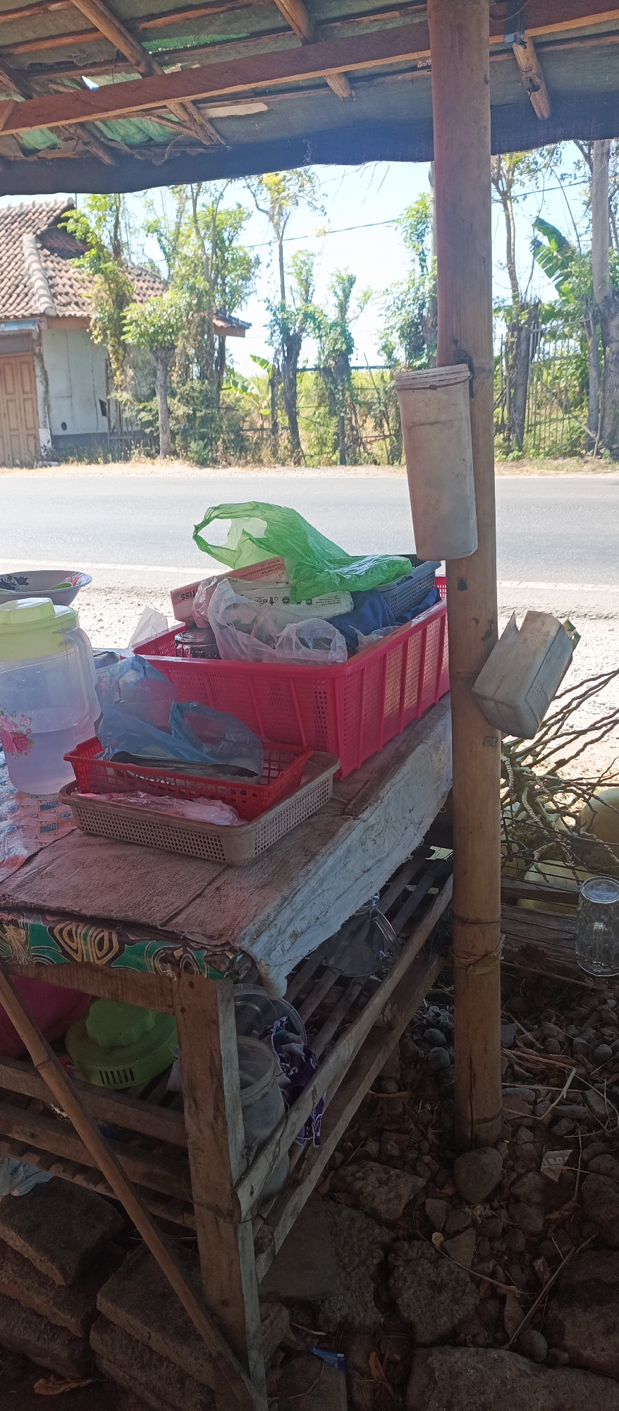 In Jatisari, Indonesia, this image captures a street-side vendor stall under a makeshift shelter with a corrugated metal roof supported by wooden beams. In the foreground, a wooden table holds various items, including plastic baskets filled with goods, a large plastic water jug, and several plastic bags. A prominent red plastic basket contains a green plastic bag and other items. To the right, a large wooden pole supports two repurposed plastic containers, one at the top and another smaller one below. The ground is covered with dirt, stones, and debris. In the background, a road runs parallel to the stall, with trees and a house visible beyond the road, suggesting a rural or semi-rural setting. The lighting indicates it is daytime.