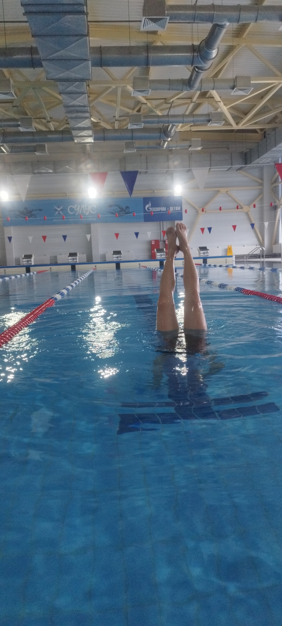 The image shows a person performing a handstand in a swimming pool. Only the person's legs and feet are visible above the water's surface. The pool is located indoors, likely in an aquatic center, with starting blocks, lane markers, and flags above the surface of the water. A banner in the background reads "ГАЗПРОМ - ДЕТЯМ", suggesting a connection with Gazprom. The location is Yakutsk, Russia. The pool bottom has blue tiles. The water is clear enough to see a vague shadow of the person performing the handstand.
