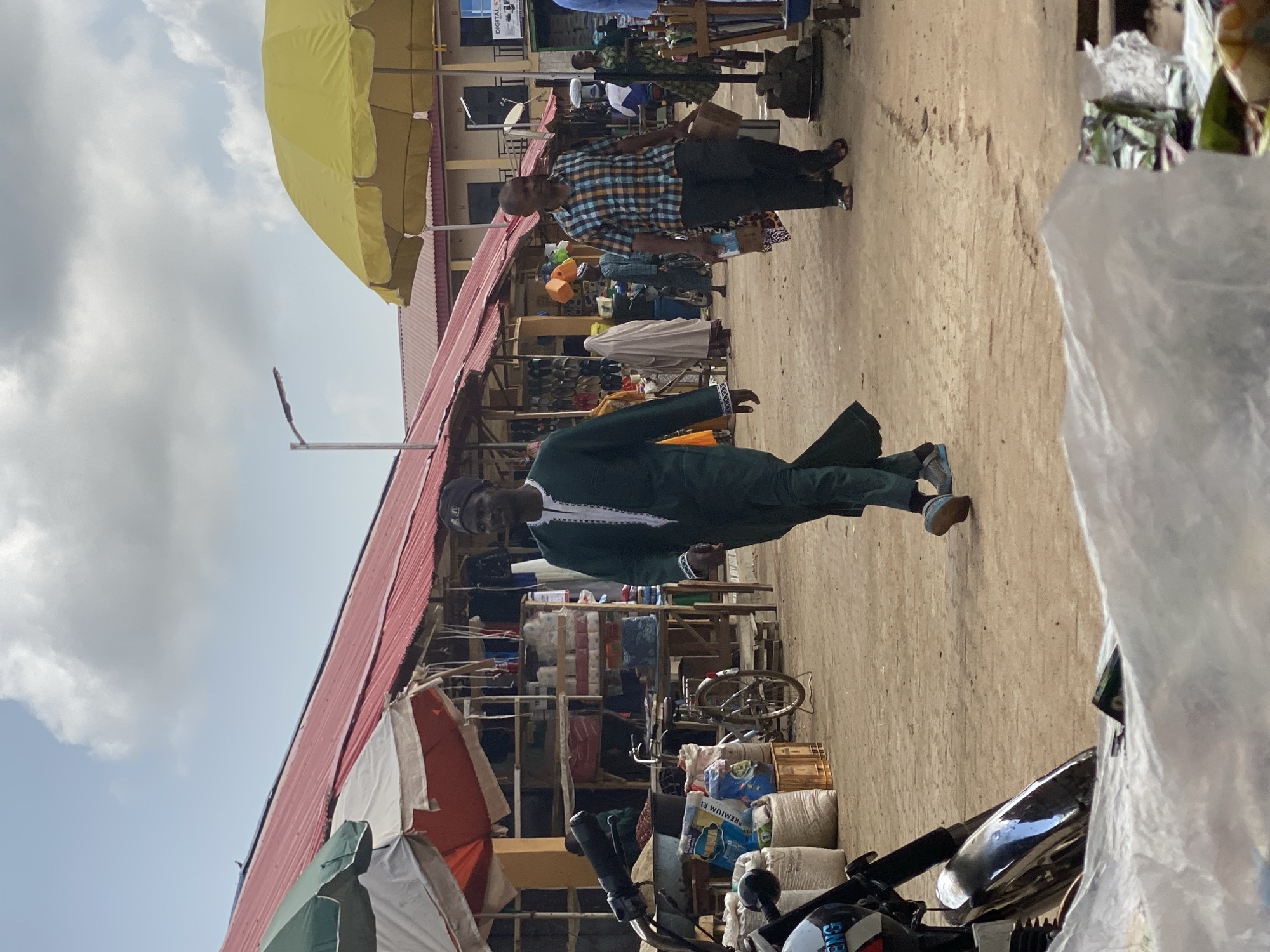 A man in a dark green robe and cap walks in front of shops under a red-roofed structure, lined with various items for sale, at what appears to be an open-air market in Kafanchan B, Nigeria. Another man in a plaid shirt walks in the background. There is a motorcycle parked in the foreground, partially covered by a white plastic sheet. Other items for sale include bags of goods and assorted household items. A yellow umbrella provides shade to the market area. The sky above is partly cloudy.