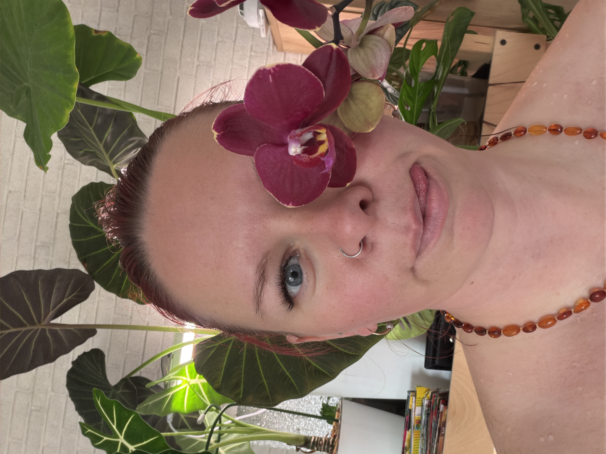 A close-up, slightly tilted portrait of a person with pale skin and blue eyes is framed by greenery and a red orchid placed over their forehead. The person has a silver nose ring and is wearing a beaded amber necklace. The background features various houseplants with large green leaves, as well as a light-colored wooden shelf and a glimpse of a white brick wall. The presence of houseplants and the interior setting suggest an indoor location. There is no information to suggest this photograph was taken in Chaikovsky, Russia.