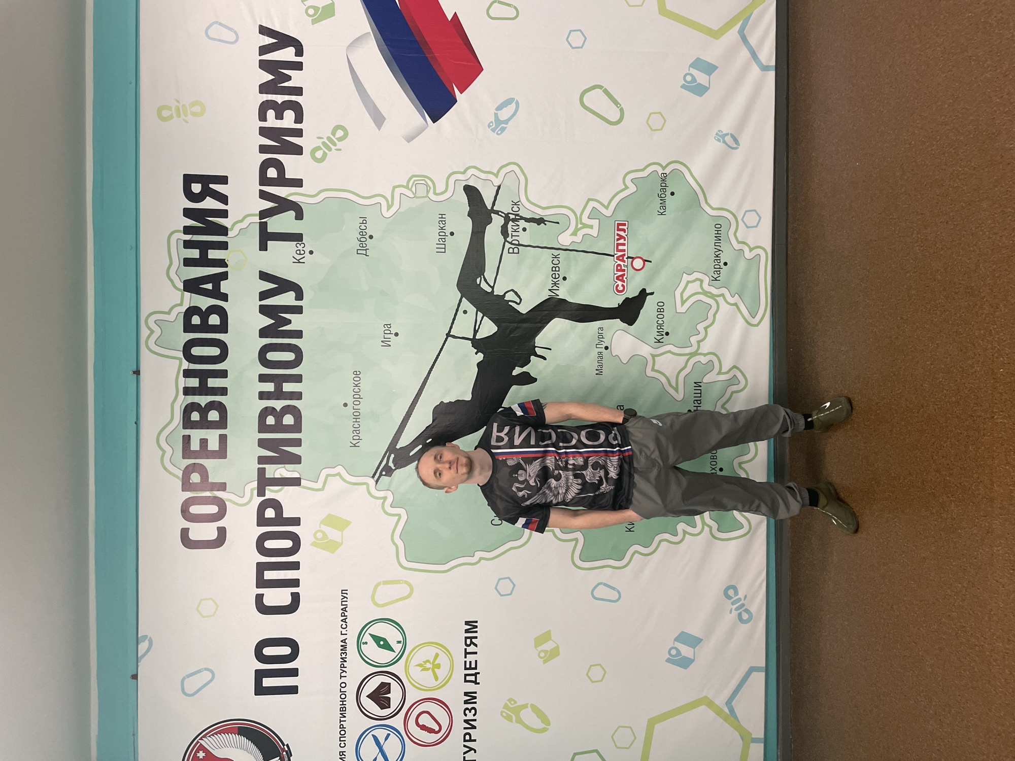 A man stands indoors, posing for a photograph in front of a large vertical banner. He looks directly at the camera with a neutral expression. The man is wearing a dark short-sleeved t-shirt adorned with a detailed graphic of a double-headed eagle and the word "РОССИЯ" (Russia), gray cargo pants, and olive-green athletic shoes.
The banner behind him features a map, likely of the Udmurt Republic in Russia, with various cities marked, including "Ижевск" (Izhevsk), "Воткинск" (Votkinsk), "Дебесы" (Debesy), and "Камбарка" (Kambarka). The city "САРАПУЛ" (Sarapul) is highlighted in red on the map. A large silhouette of a person engaged in rappelling or climbing is centrally placed on the map, connected to Sarapul by a line. Above the silhouette, a Russian flag is unfurling.
Prominent text in Russian on the banner reads "СОРЕВНОВАНИЯ ПО СПОРТИВНОМУ ТУРИЗМУ" (Competitions in Sports Tourism) at the top and "ТУРИЗМ ДЕТЯМ" (Tourism for Children) at the bottom. Various icons related to outdoor activities, such as a compass, a tent, and a climbing carabiner, are scattered across the banner. The overall setting is indoors, with a brown floor and a light blue wall visible on the left side. The banner promotes sports tourism, seemingly for children, within the region.