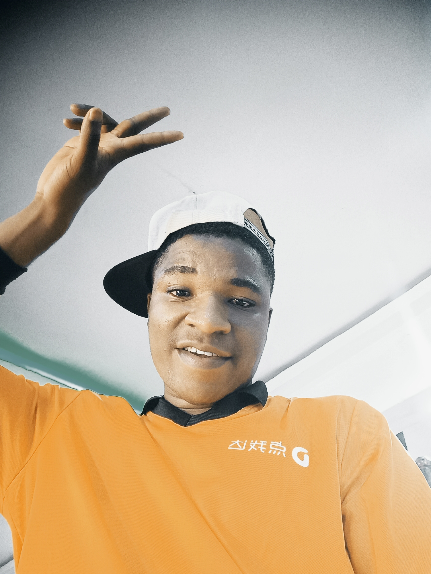 A low-angle portrait depicts a young man with brown skin, wearing a white baseball cap with a black brim and an orange t-shirt with white writing in Chinese on the left side. He smiles, looking directly at the camera, with his left arm raised and hand open, fingers extended. The background appears to be a white ceiling. The picture location is Bukuru, Nigeria.