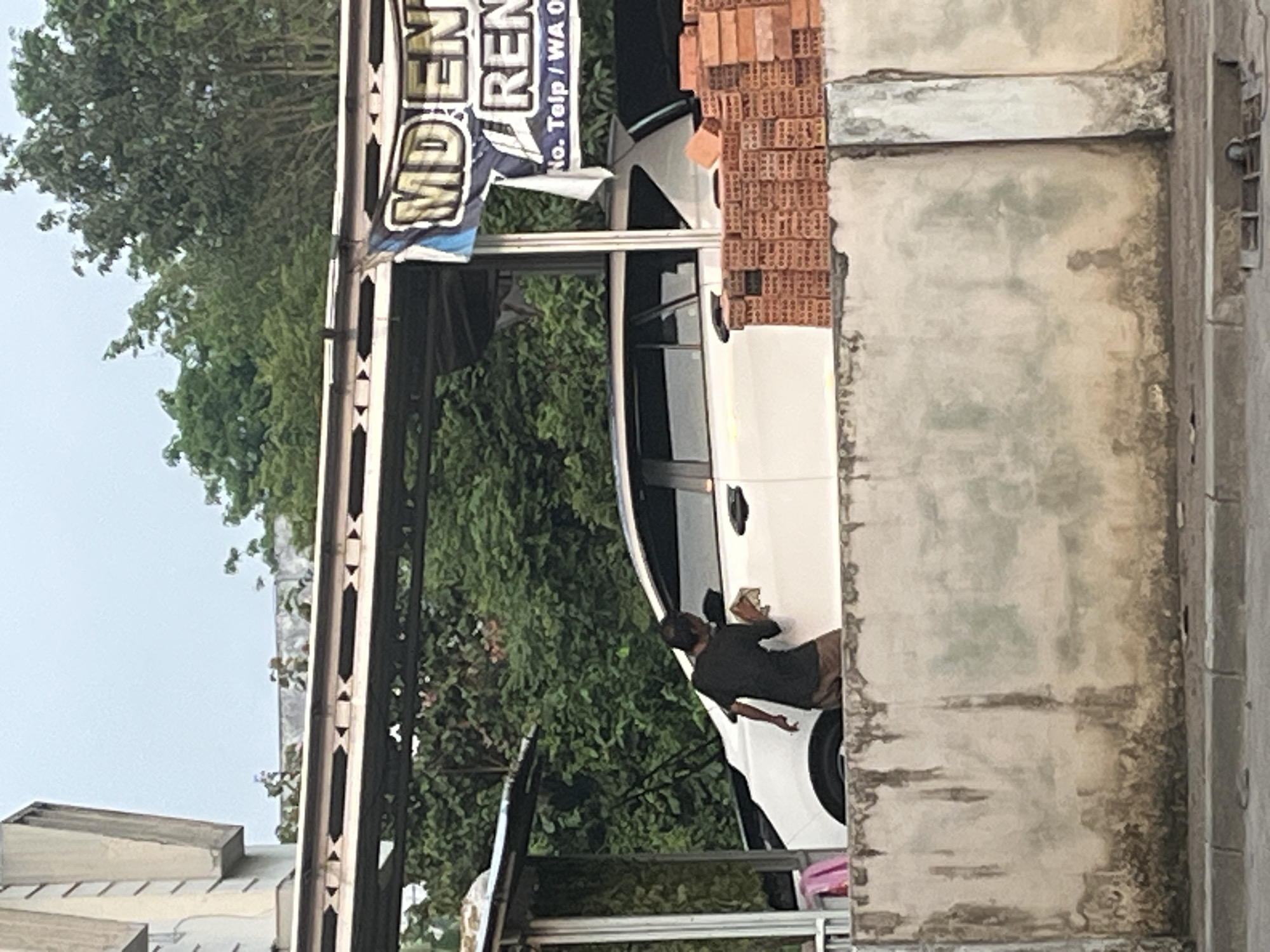 A man is seen washing a white car in front of a building with a blue and white banner that reads "MDEN RENT" and "No. Telp / WA 0". The scene appears to be an outdoor establishment in Sekayu, Indonesia, possibly a car rental or car wash. There is a stack of bricks in the foreground. The environment is somewhat overgrown with trees and foliage in the background. The sky is overcast.