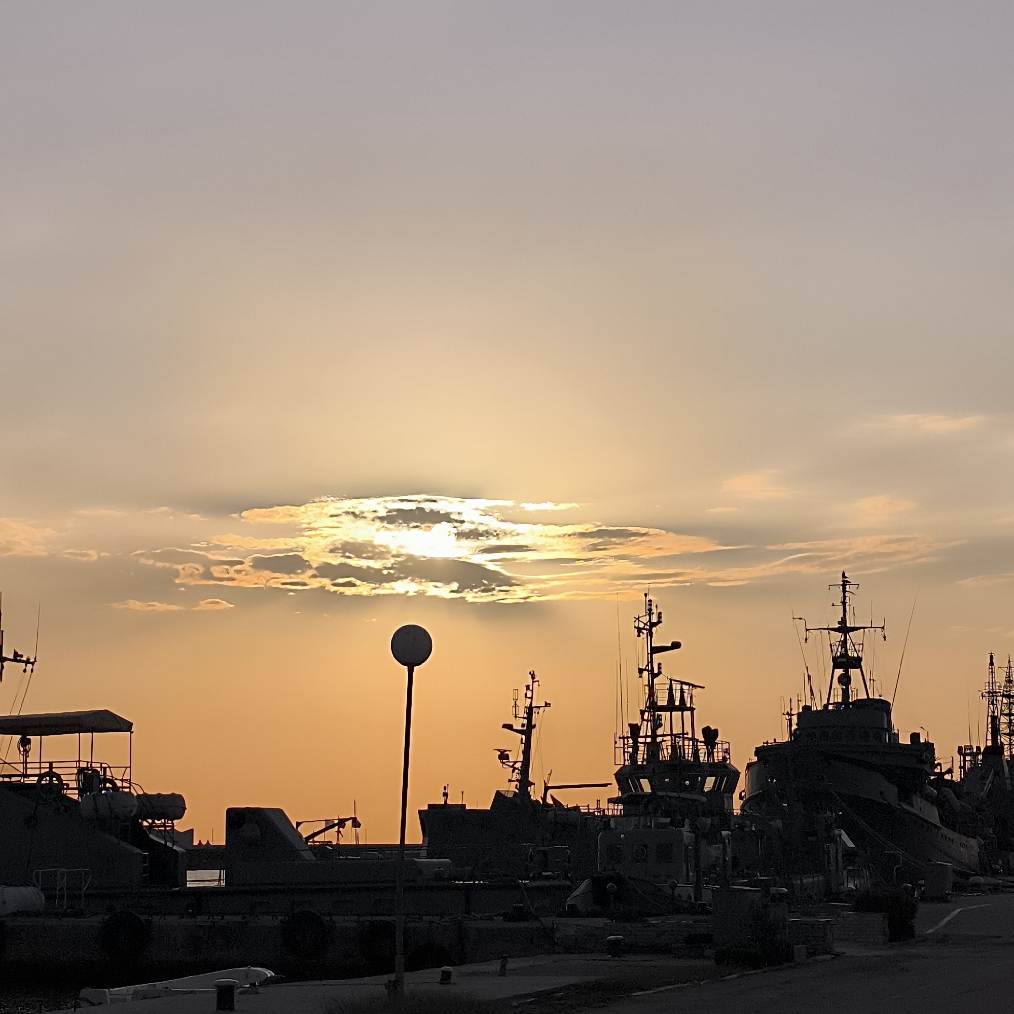 This is a photograph of the Varna Port in Varna, Bulgaria, taken at dusk. The sky is a gradient of soft grays and oranges, with a bright sun partially obscured by scattered clouds. Several ships are docked in the foreground, silhouetted against the twilight sky. The ships appear to be naval vessels or cargo ships, with their masts and superstructures clearly visible. A solitary lamppost stands in the middle ground, adding a vertical element to the scene. The overall mood is calm and serene, capturing the quiet beauty of the port as day turns into night.