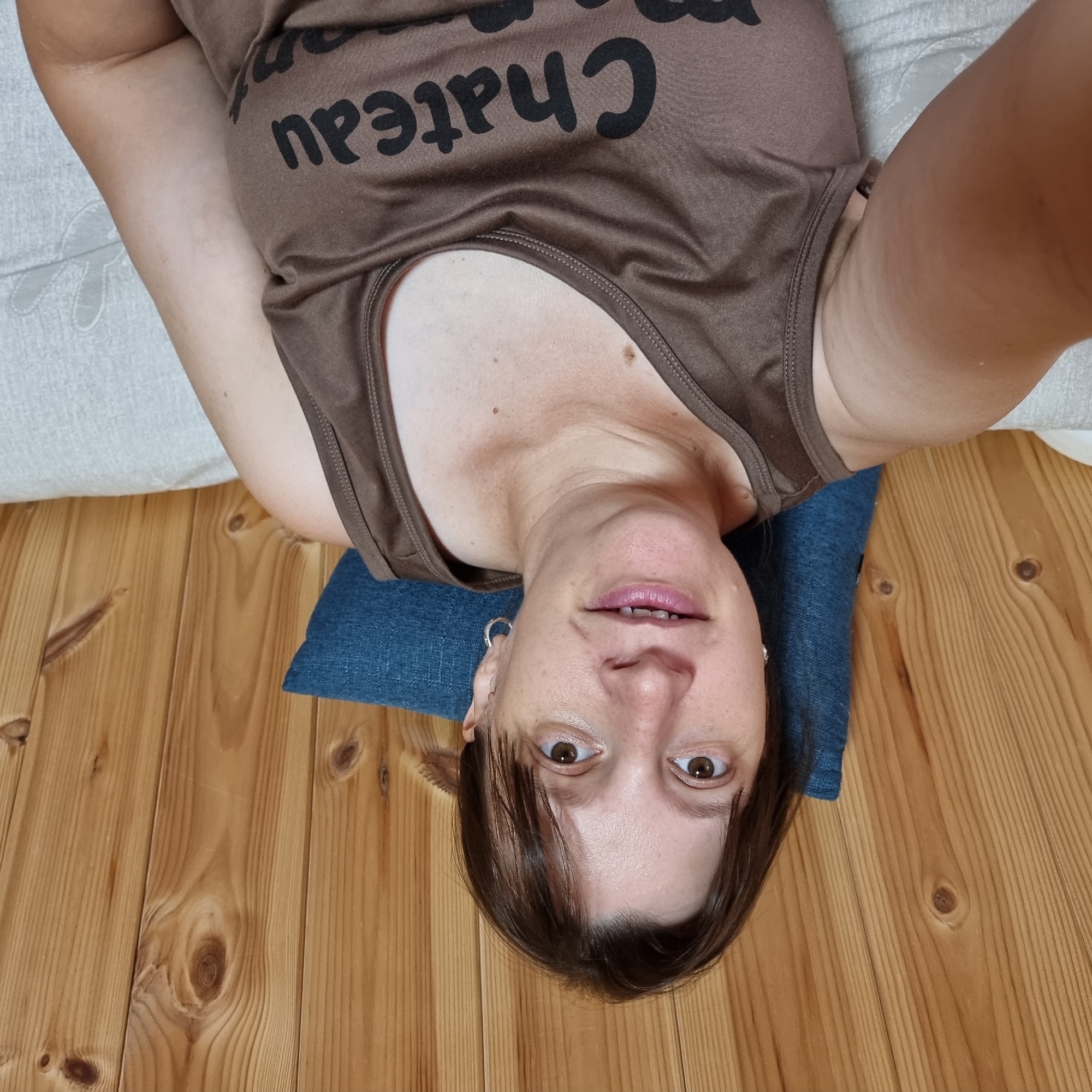 A woman is taking a selfie in a room with wood-paneled walls. She is wearing a brown tank top with black lettering that appears to say "Chateau" and "mon." A blue pillow is behind her. She has brown hair and brown eyes and is wearing a silver hoop earring in her visible ear. The woman is resting on a white, textured surface. The image does not provide information that would allow for the determination of the location in Dunnae-myeon, South Korea.