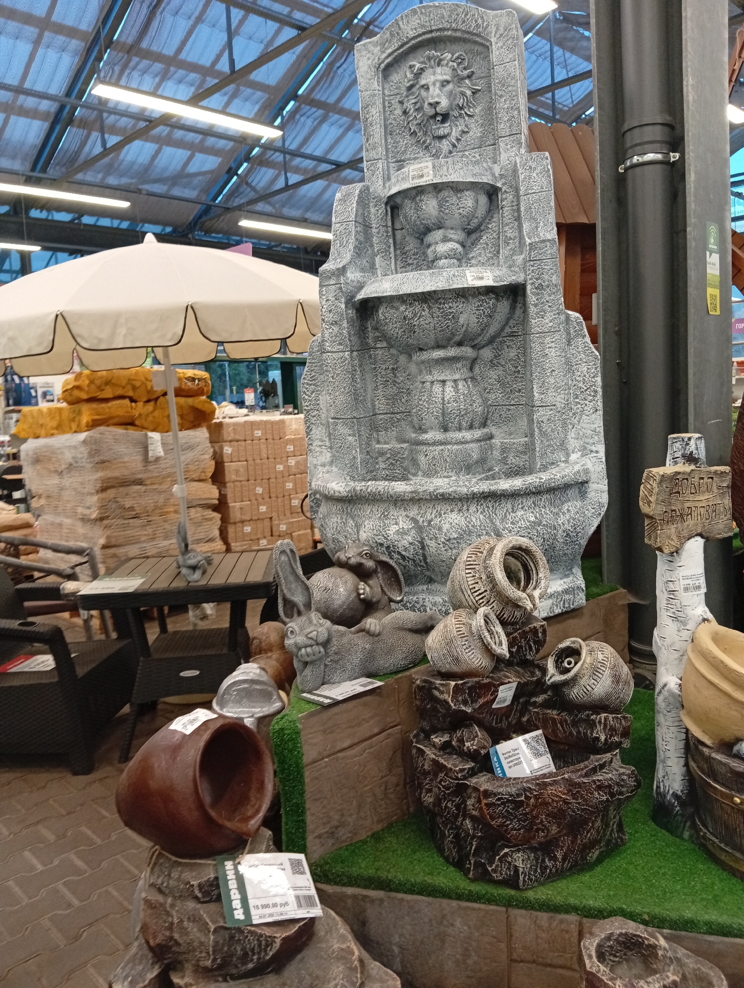 The image depicts an indoor retail environment, likely a garden or home decor store, with various decorative items displayed. In the foreground, a large, grey, stone-like fountain features a lion's head with water spouts, and two tiered basins. To the right of the fountain, a smaller, multi-tiered rock formation with several decorative pots is placed on artificial grass. Further right, a white signpost resembling a birch tree trunk with carved text reads "ДОБРО ПОЖАЛОВАТЬ" (Welcome). To the left of the fountain, a large brown ceramic pot rests on a rock. Behind the fountain and pots, stacks of packaged merchandise are visible, including what appear to be paving stones or tiles. A large white patio umbrella is situated behind the stacks of merchandise. In the background, the store's ceiling structure with exposed beams and fluorescent lighting is visible. The setting appears to be a retail space, possibly a large hardware or garden supply store. There are no people clearly visible in the image. The overall environment is organized for product display. While the city of Pushkino, Russia is mentioned, there are no specific visual cues within the image to confirm this exact location, other than potential signage in Russian.