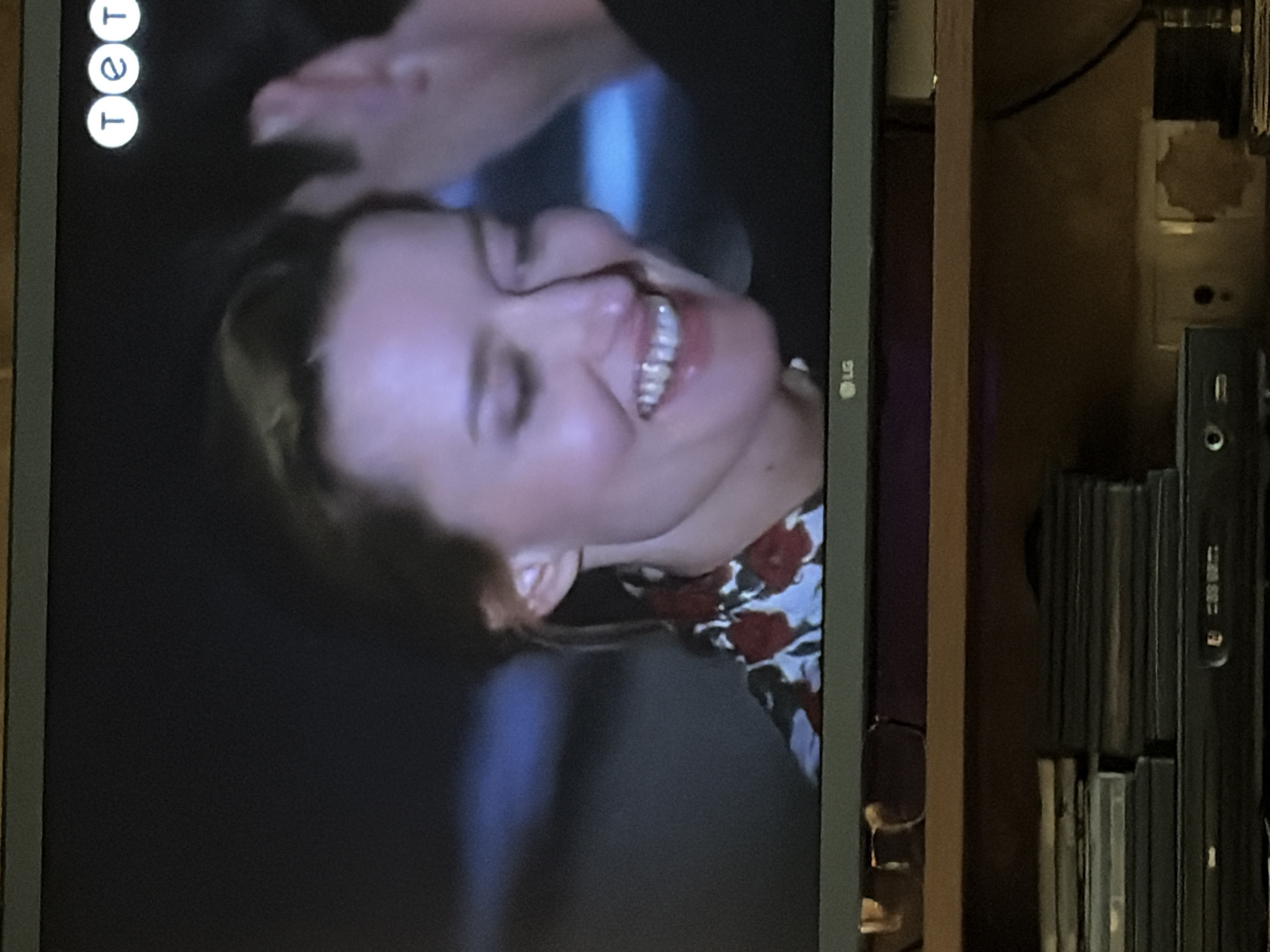 A television screen displays a close-up shot of a woman with blonde hair, wearing a floral print top with red and white flowers. She is smiling with her eyes closed and her head tilted back. Her teeth are visible. A hand is touching her hair from the right side of the frame. In the upper right corner of the screen, a logo with three white circles containing the letters "T", "e", and "T" is visible. In the lower center of the screen, an "LG" logo is present. The background is dark and out of focus, with some blue lighting visible. Below the television, a wooden shelf holds stacks of DVDs or similar media and a black electronic device with buttons and ports. The environment appears to be indoors, likely a living room or entertainment area.