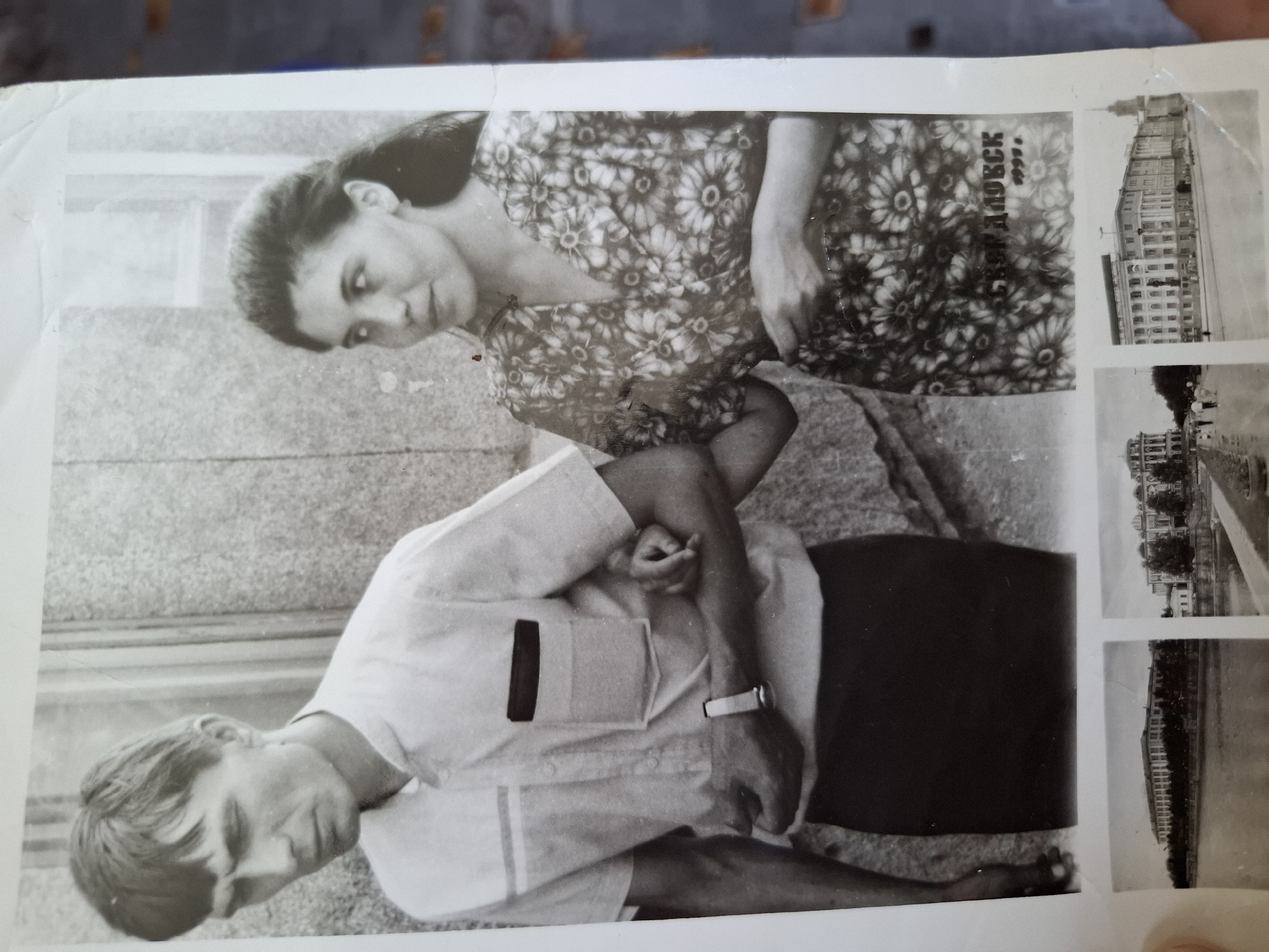 The image shows a black and white photograph with two people, a man and a woman, posing. The man is in a light-colored shirt, wearing a watch. The woman is in a dark floral dress. There are other smaller photographs adjacent to the man and woman on the right side of the photograph. One shows a large building and another shows the same building from a distance. There is text near the woman that reads “Амзя 1999.” The image is from Амзя, Russia.