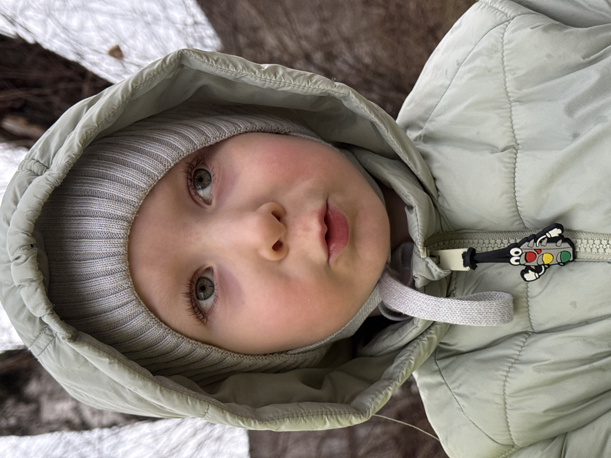 A close-up, slightly angled, eye-level shot depicts a young child in a light gray hooded jacket, with a matching gray knit cap visible under the hood. The child's face is the primary focus, displaying fair skin with rosy cheeks and a slightly open mouth. The jacket's zipper is visible on the right side, adorned with a colorful traffic light charm. In the periphery, blurred segments of what appears to be snow and wooden textures are present. There is no verifiable information to place this image in Perm, Russia.