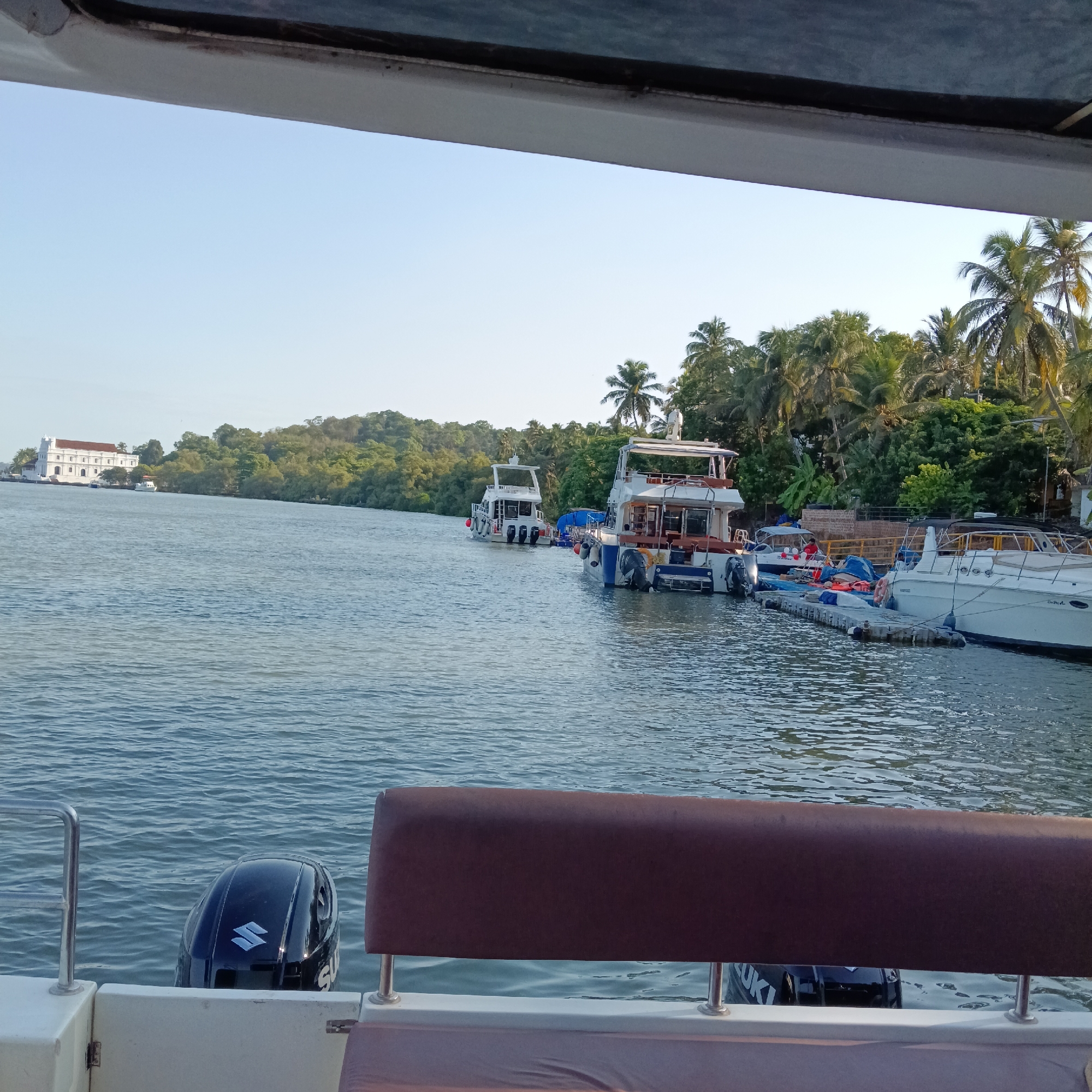 The image depicts a waterfront scene in Britona, India. The view is from a boat, looking out towards the Mandovi River. Several boats are docked along the waterfront, including a white yacht and larger vessels with multiple decks. On the opposite bank, a large, white colonial-style building stands amidst lush greenery and palm trees. The foreground shows the back of the boat the photo was taken from, including a brown cushioned seat and a black Suzuki outboard motor. The sky is clear and blue, suggesting a bright day.