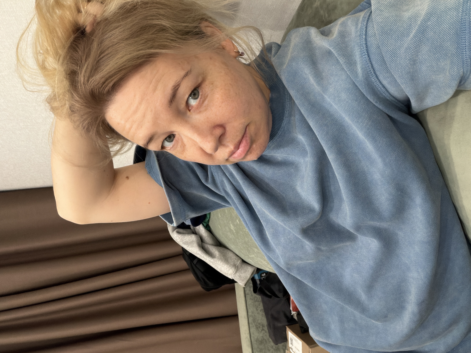 A blonde woman with her hair in a bun is wearing a light blue shirt, looking into the camera. She is holding her hair up with her right hand. The background includes brown curtains, a gray sofa, and assorted clothing items. There is no information in the image to indicate that the location is Izhevsk, Russia.