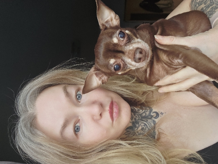 A close-up selfie shows a person holding a small dog, with both subjects looking towards the camera. The image is taken at a slight angle, with the person's head tilted.
The person has fair skin, striking light blue eyes, and long, straight blonde hair that appears to have some grey or white strands mixed in. Her eyebrows are light, matching her hair color. Her lips are full, slightly parted, and tinted a natural pinkish red. A prominent and intricate tattoo covers her neck and extends onto her upper chest/shoulder area. The tattoo is done in blackwork style, featuring ornate, possibly floral or lace-like patterns, with a clear depiction of a crescent moon and stars towards the center of her collarbone. Her bare shoulder or upper chest is visible, suggesting a strapless garment or partial nudity.
She is holding a small dog, likely a Chihuahua or similar toy breed. The dog has short, shiny brown fur, with lighter, almost tan markings around its muzzle, chin, and above its eyes. Its ears are large, upright, and pointed. The dog's eyes are dark, round, and reflective, also looking directly at the camera. It is nestled against the person's upper body, held securely by her hands.
The background is dark and out of focus, providing a soft contrast to the subjects and suggesting an indoor setting with limited lighting or a plain dark wall. The overall impression is one of affection and companionship between the person and their pet. There is no visible text in the image.